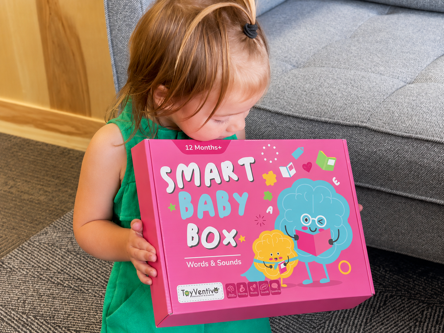 Menina com cabelo castanho-avermelhado segurando uma caixa rosa "Smart Baby Box" com ilustrações de personagens fofos, uma com óculos leyendo um livro, e a outra segurando um celular, ao lado de um sofá cinza em ambiente interno.