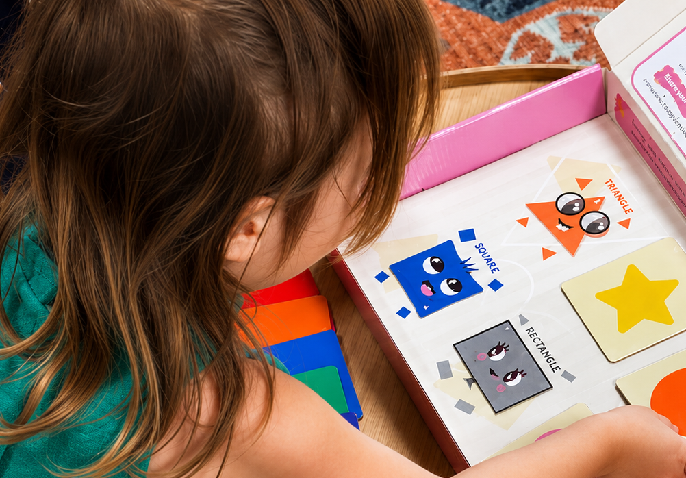 menina brincando com cartas de formas geométricas coloridas, incluindo triângulo, quadrado e retângulo, com desenhos fofos e olhos, em uma caixa de jogo