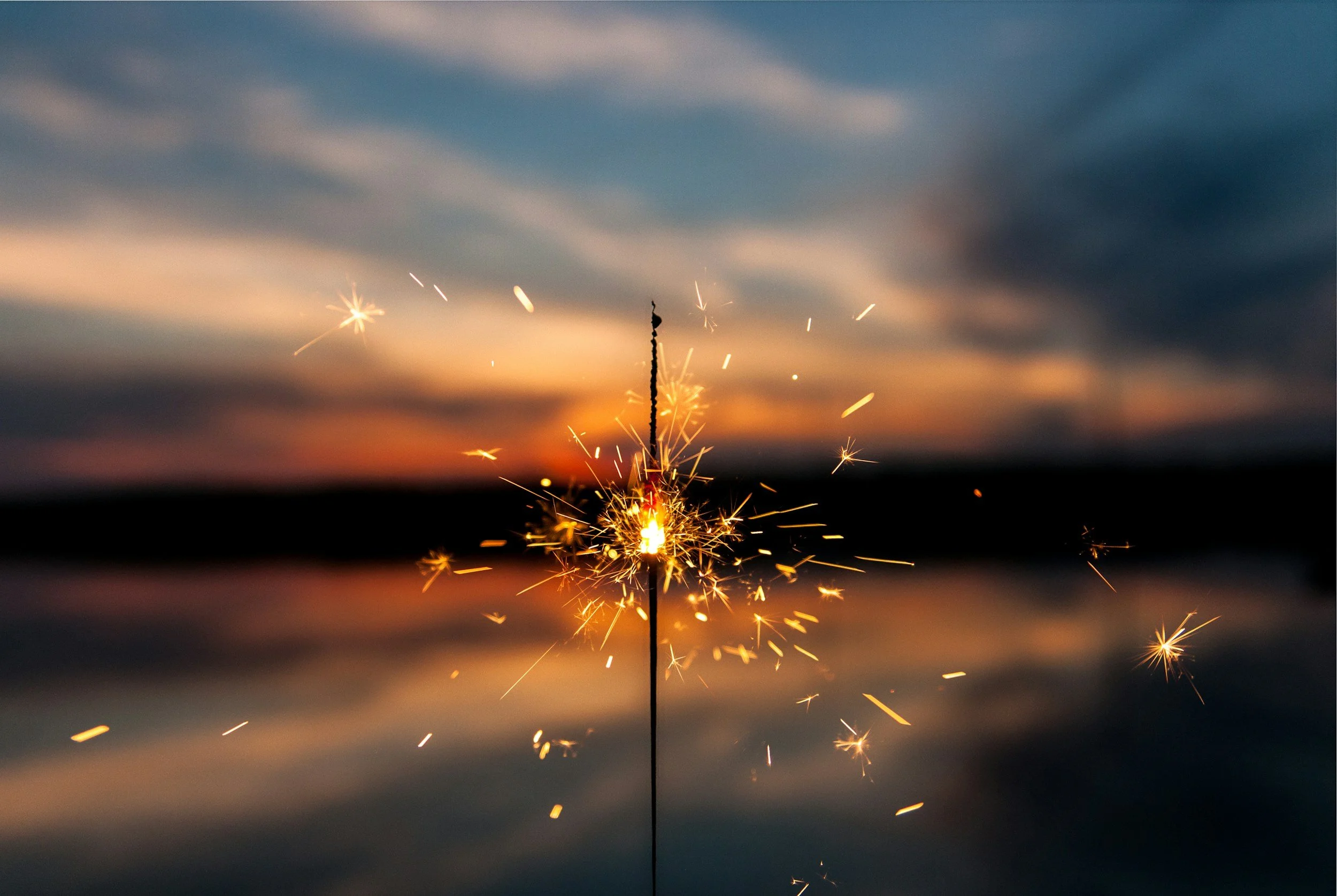 A sparkler firework burning with bright sparks against a twilight sky, with a dark horizon and clouds overhead.