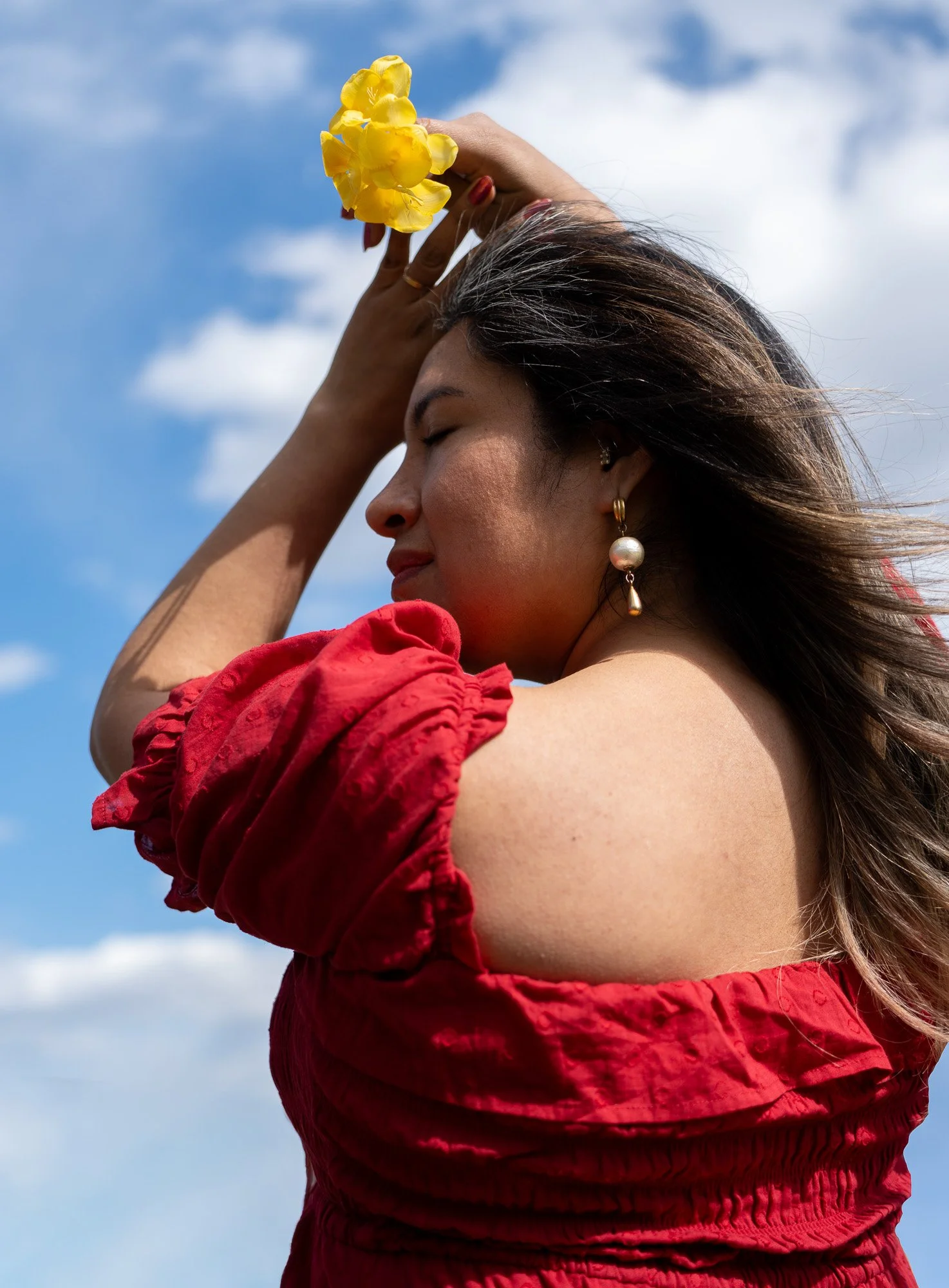 A woman with long dark hair, closed eyes, and wearing pearl earrings, is holding a yellow flower above her head against a blue sky with some clouds. She is wearing a red ruffled dress.