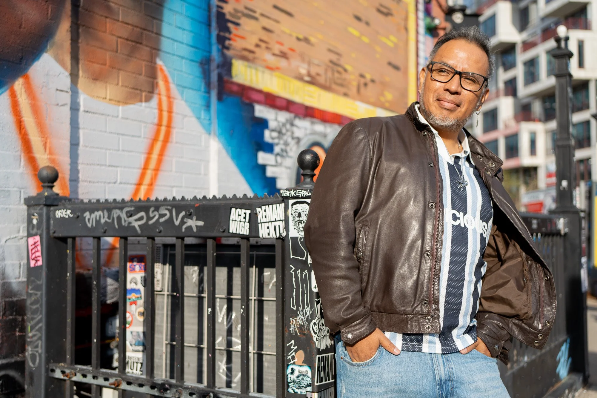 A man wearing glasses, a leather jacket, and a striped shirt, standing in front of a colorful urban mural on a brick wall, with a city street and apartment buildings in the background.