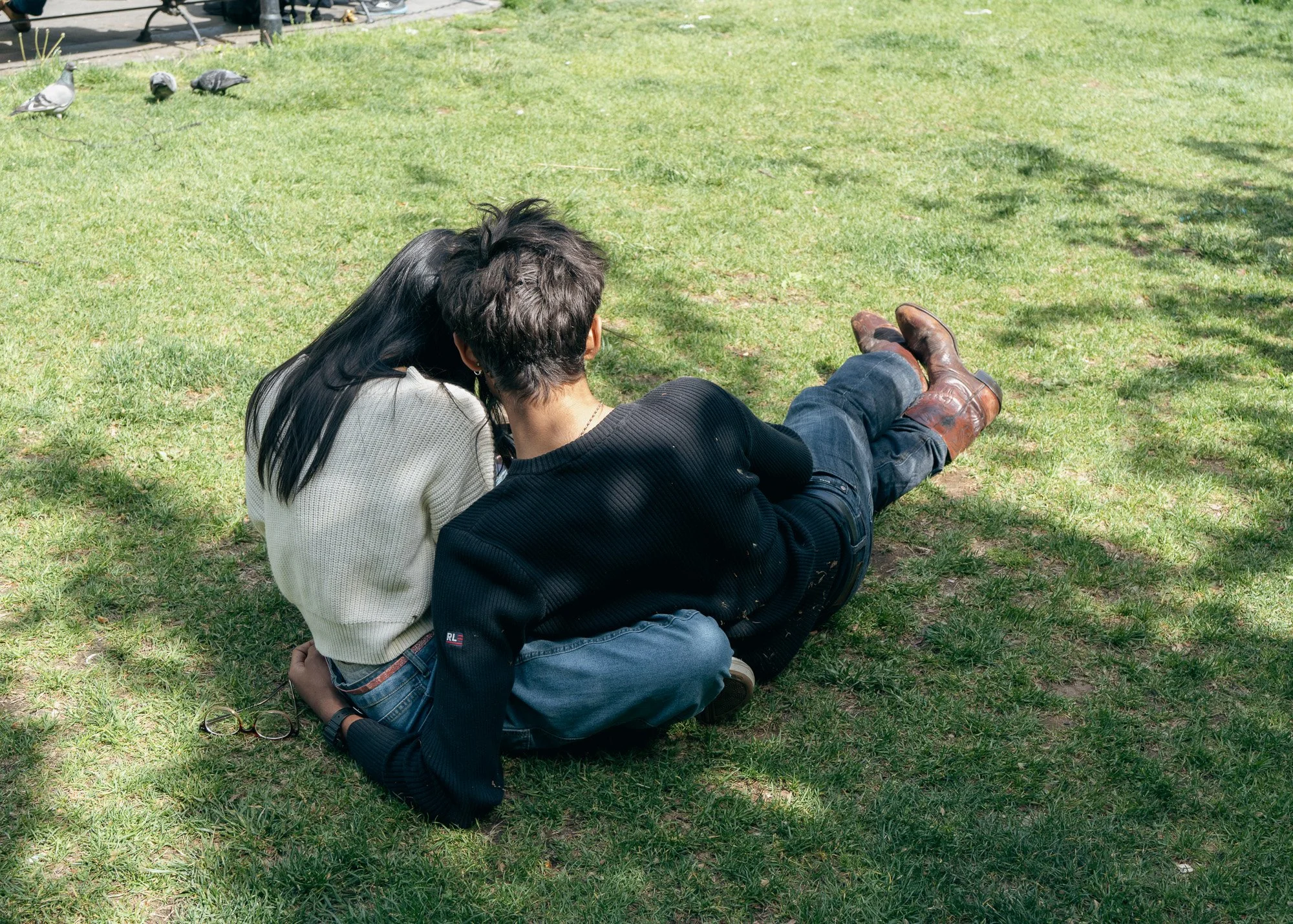 A man and woman with dark hair sitting on the grass in a park, facing away from the camera. The man is wearing a black shirt, jeans, and brown boots, while the woman is wearing a white sweater and blue jeans. In the background, there are pigeons and 