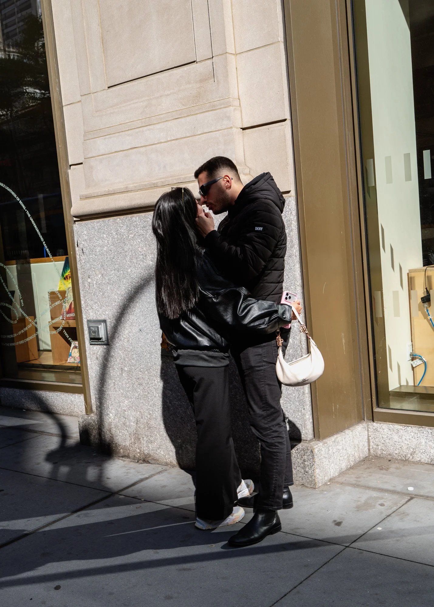 A man and woman sharing an intimate moment on the street, nose to nose, with the woman holding a phone, standing close against a building with a glass window and sidewalk.