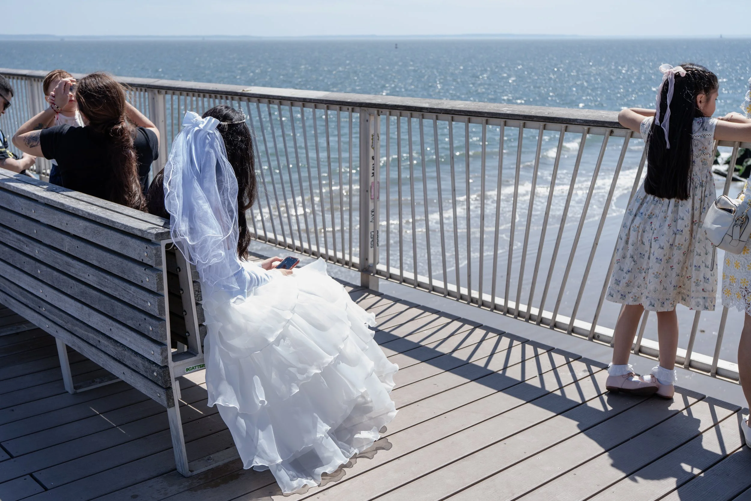 People, including a girl in a wedding dress, sitting and standing on a wooden pier overlooking the ocean, with a girl in a floral dress and skate shoes standing by the railing.