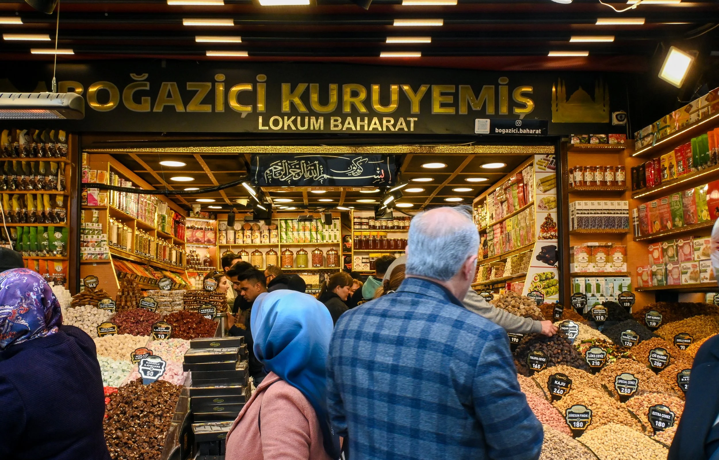 A busy market stall selling Turkish spices and dried fruit, with customers browsing and shopping.