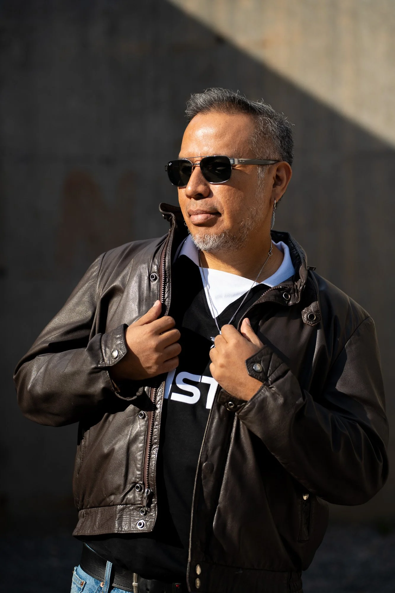 A man with gray hair, wearing black sunglasses, a black T-shirt, and a brown leather jacket, standing outdoors against a dark background.