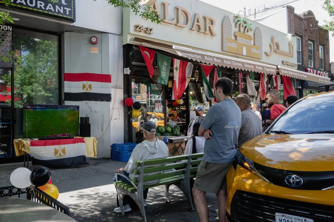 People watching a soccer game on a television outside a Mediterranean grocery store decorated with flags and balloons, with a yellow taxi and trees in the background.
