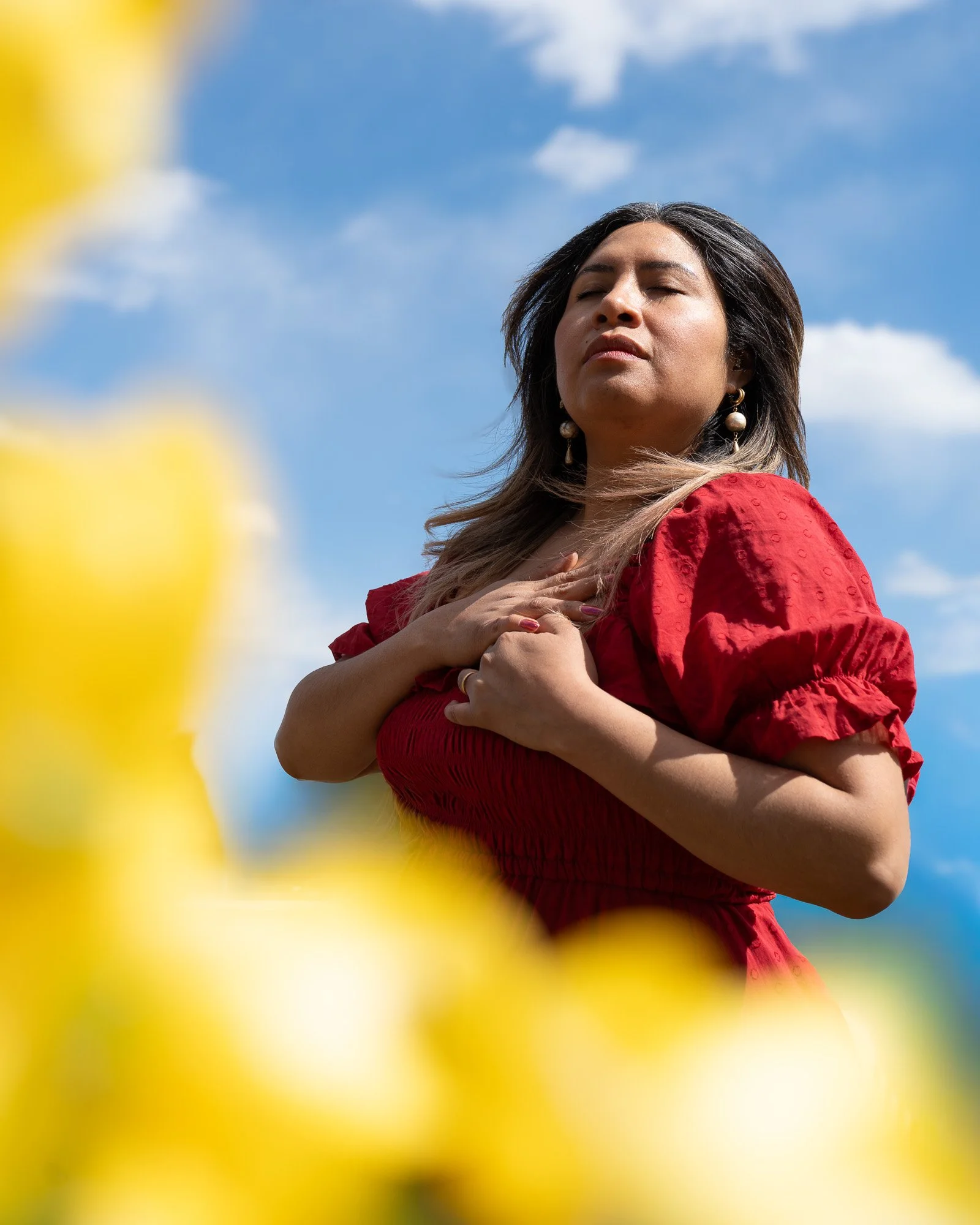 A woman with long dark hair wearing gold earrings and a red dress stands outdoors with her eyes closed and hands on her chest, against a blue sky with clouds.
