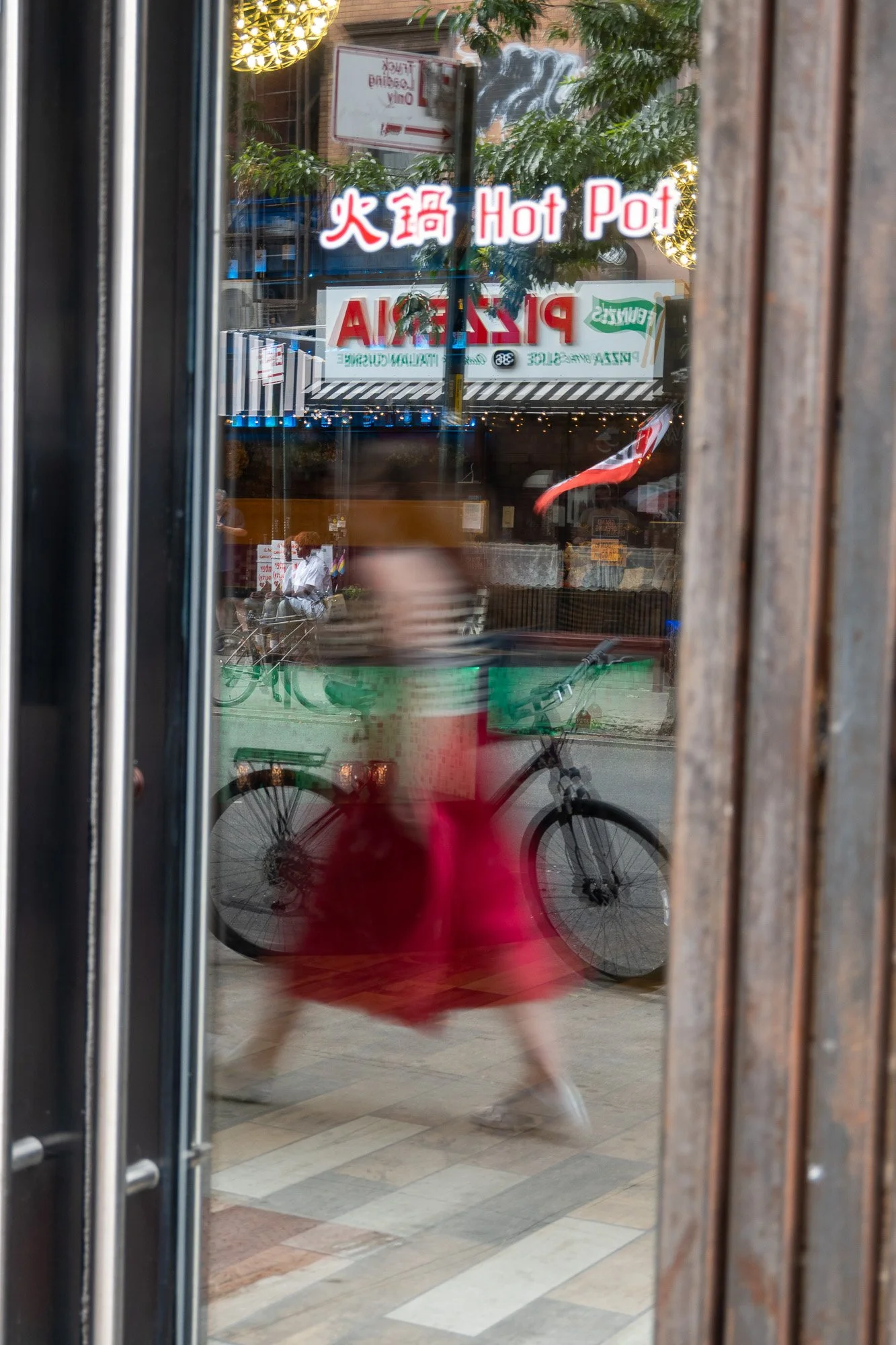 A view through a glass door showing a sign that reads 'Hot Pot' in English and Chinese characters, with the interior of a restaurant and bicycles parked outside visible in the reflection.