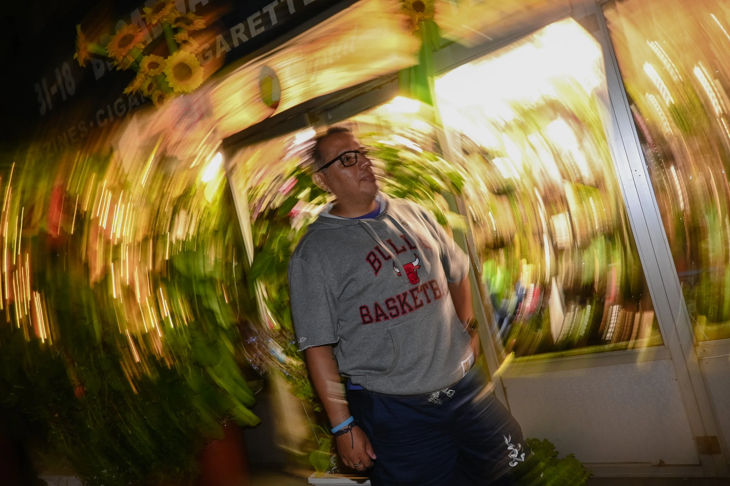 A man wearing glasses and a gray Chicago Bulls basketball hoodie standing outside at night, with vibrant yellow and pink flower decorations swirling around him due to camera motion. He is looking to his left with a neutral expression.