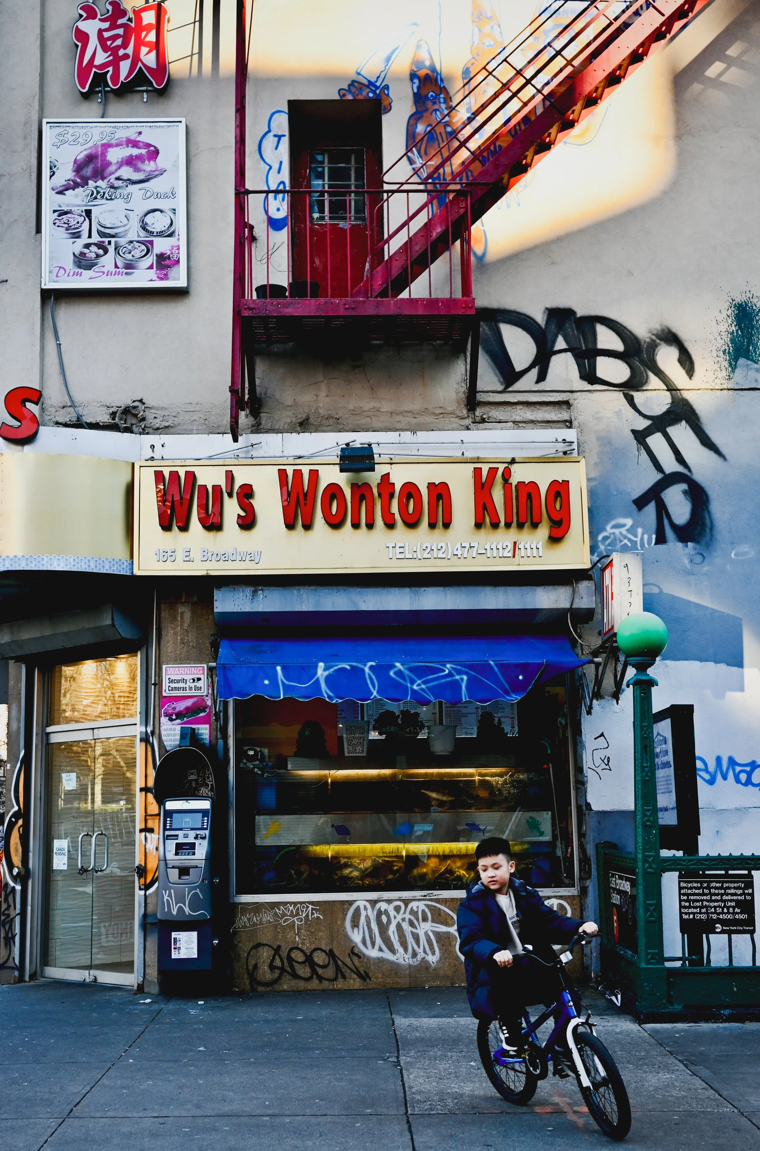 A storefront with a sign that reads 'Wu's Wonton King' and a yellow menu board below. There is graffiti on the wall and a red fire escape staircase above. A boy is riding a bicycle in front of the store.
