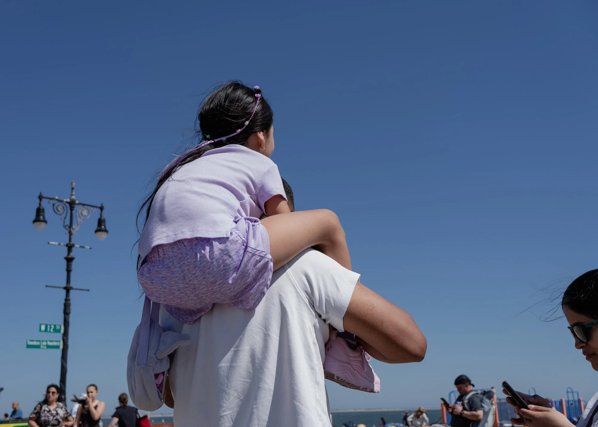 A woman carrying a young girl on her shoulders at an outdoor event on a sunny day, with a clear blue sky and people in the background.