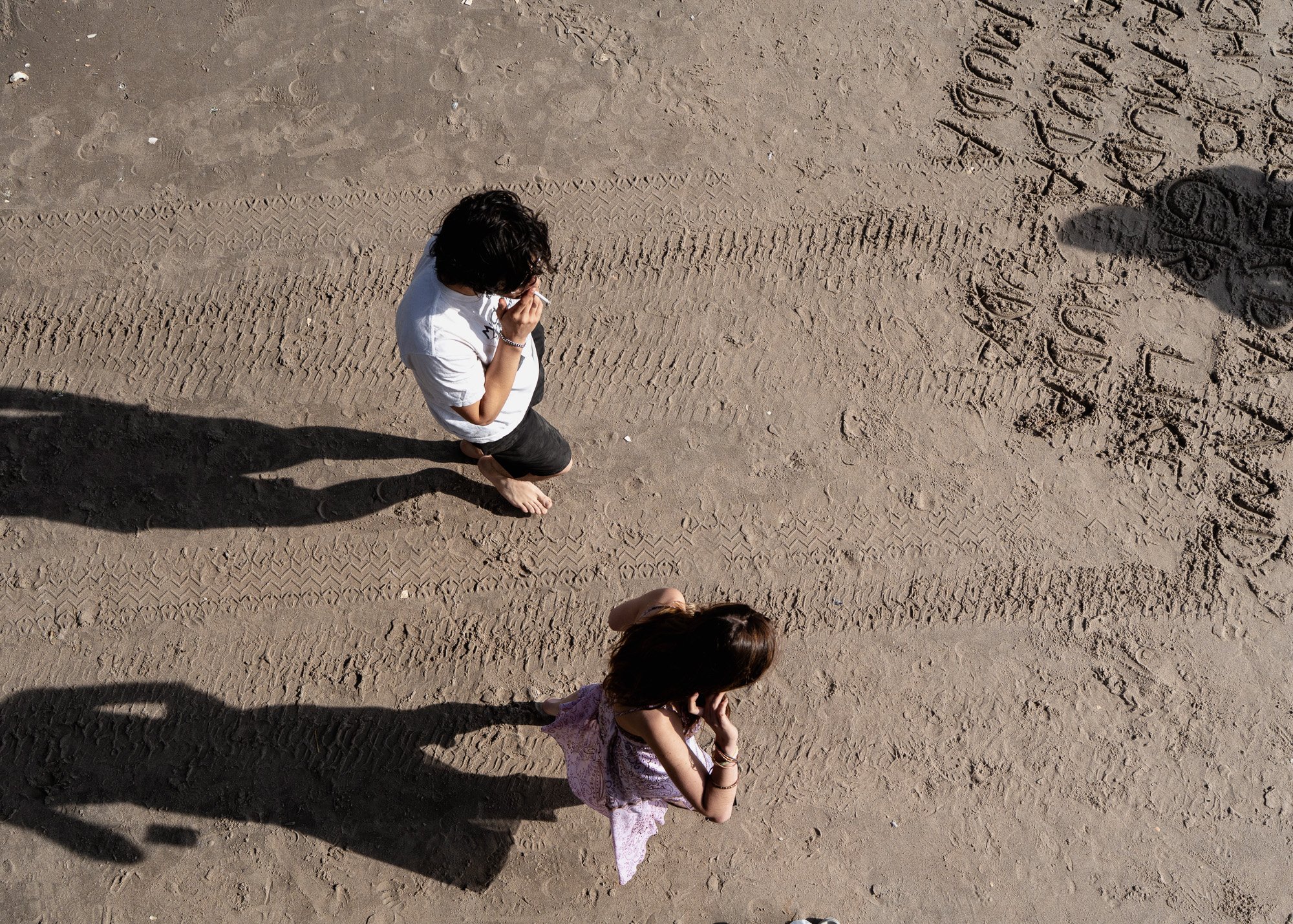 Two children standing on a dirt surface with tire tracks showing, casting long shadows and viewed from above.