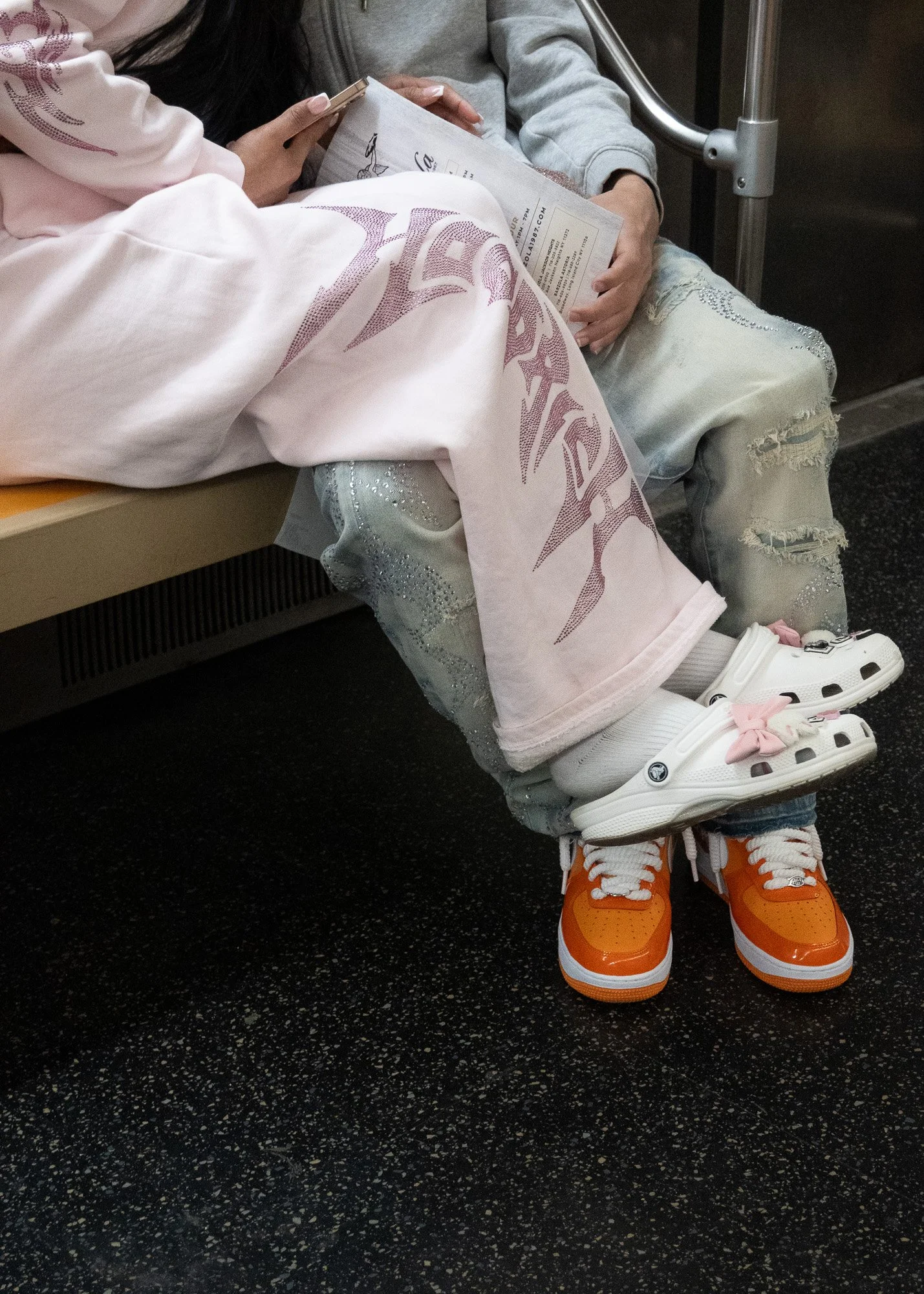 A person sitting on public transportation with legs crossed, wearing bright orange and white sneakers, white socks, distressed jeans, and a gray sweatshirt, holding a newspaper.