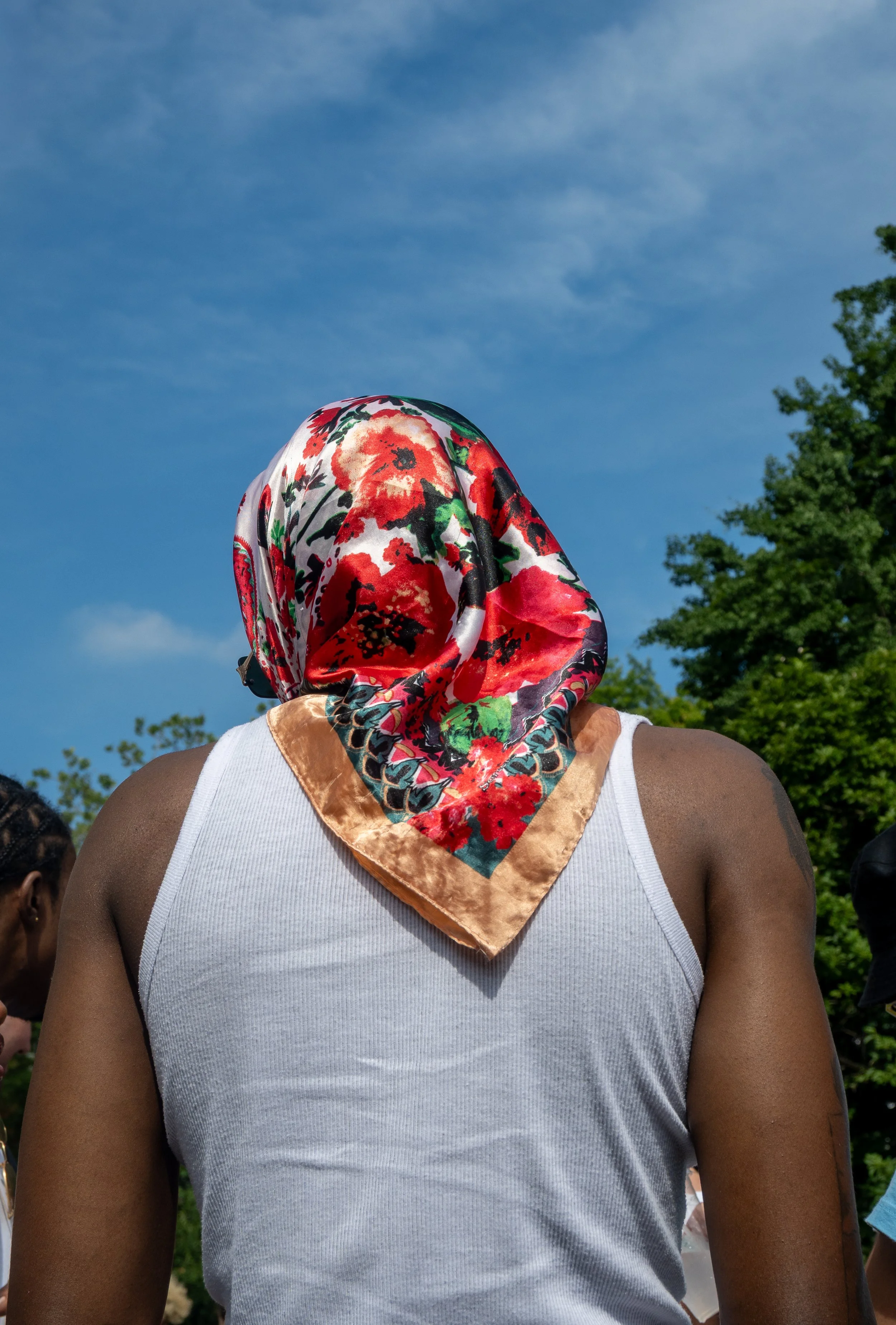 A person wearing a floral patterned headscarf and a white tank top standing outdoors on a sunny day with blue sky and green trees in the background.