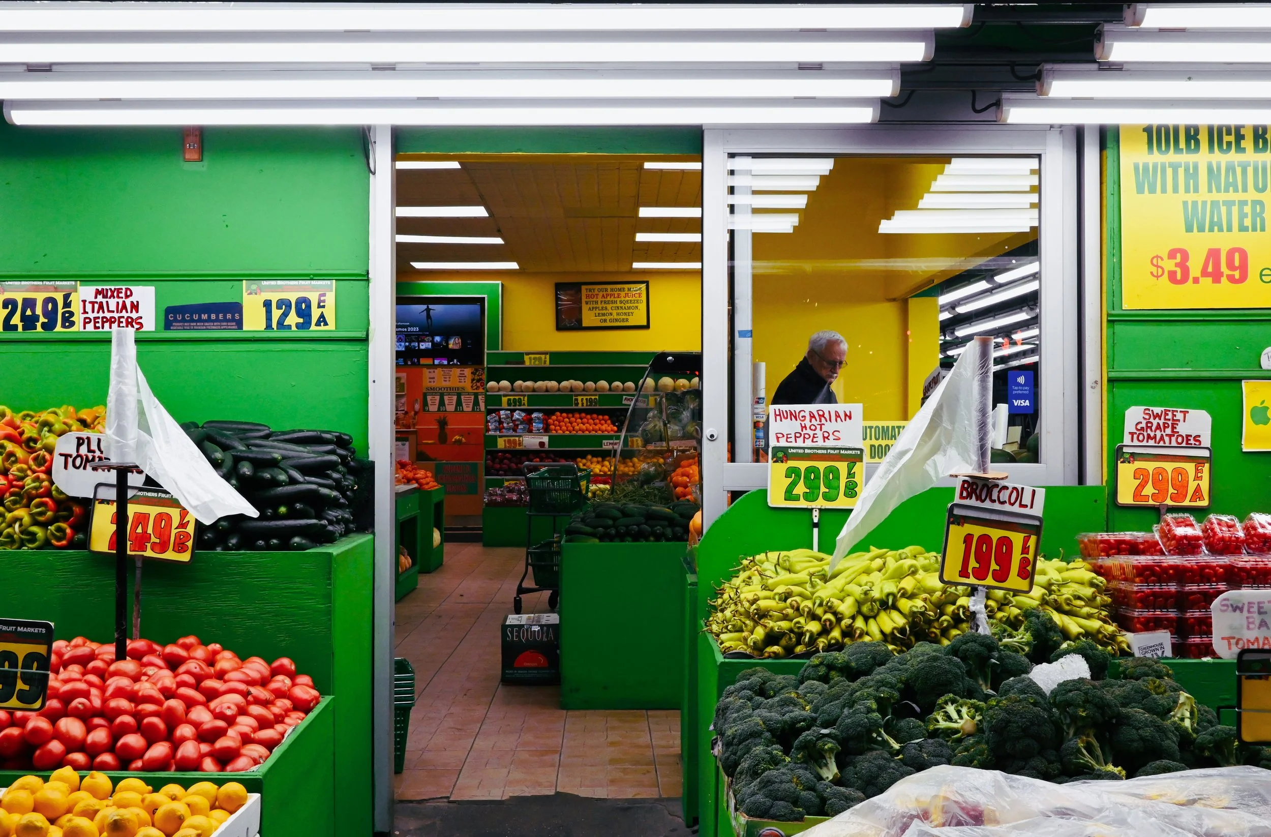 A grocery store produce section with various fruits and vegetables on display, including tomatoes, eggplants, bananas, broccoli, and peppers, with colorful price signs and a man shopping in the background.