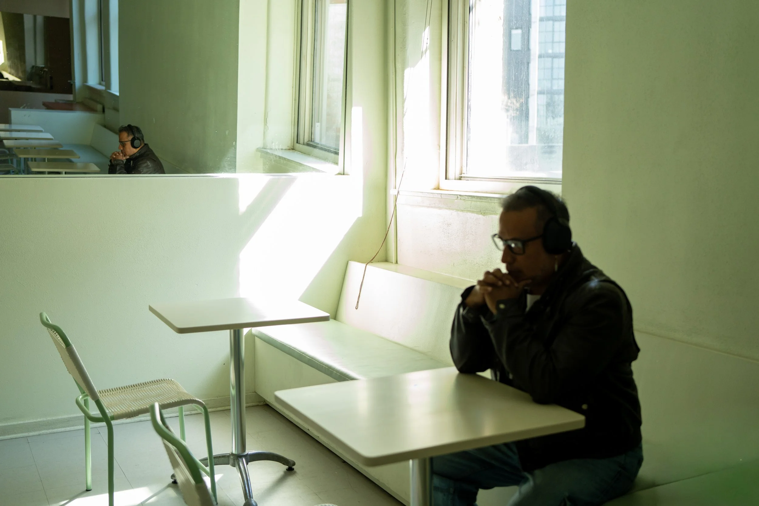 Two men wearing headphones sit alone at separate tables in a room with green walls and large windows, bathed in natural sunlight.