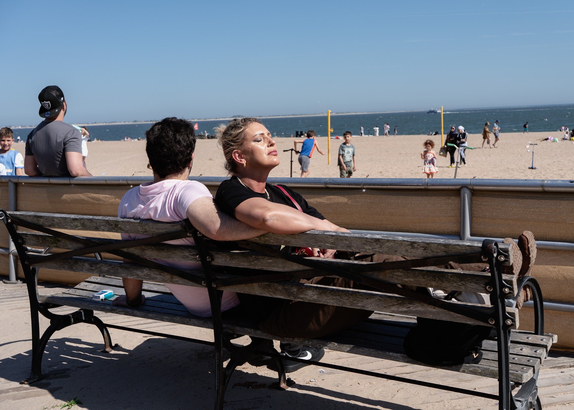 Two women sitting on a park bench near the beach with the ocean and people in the background on a sunny day.