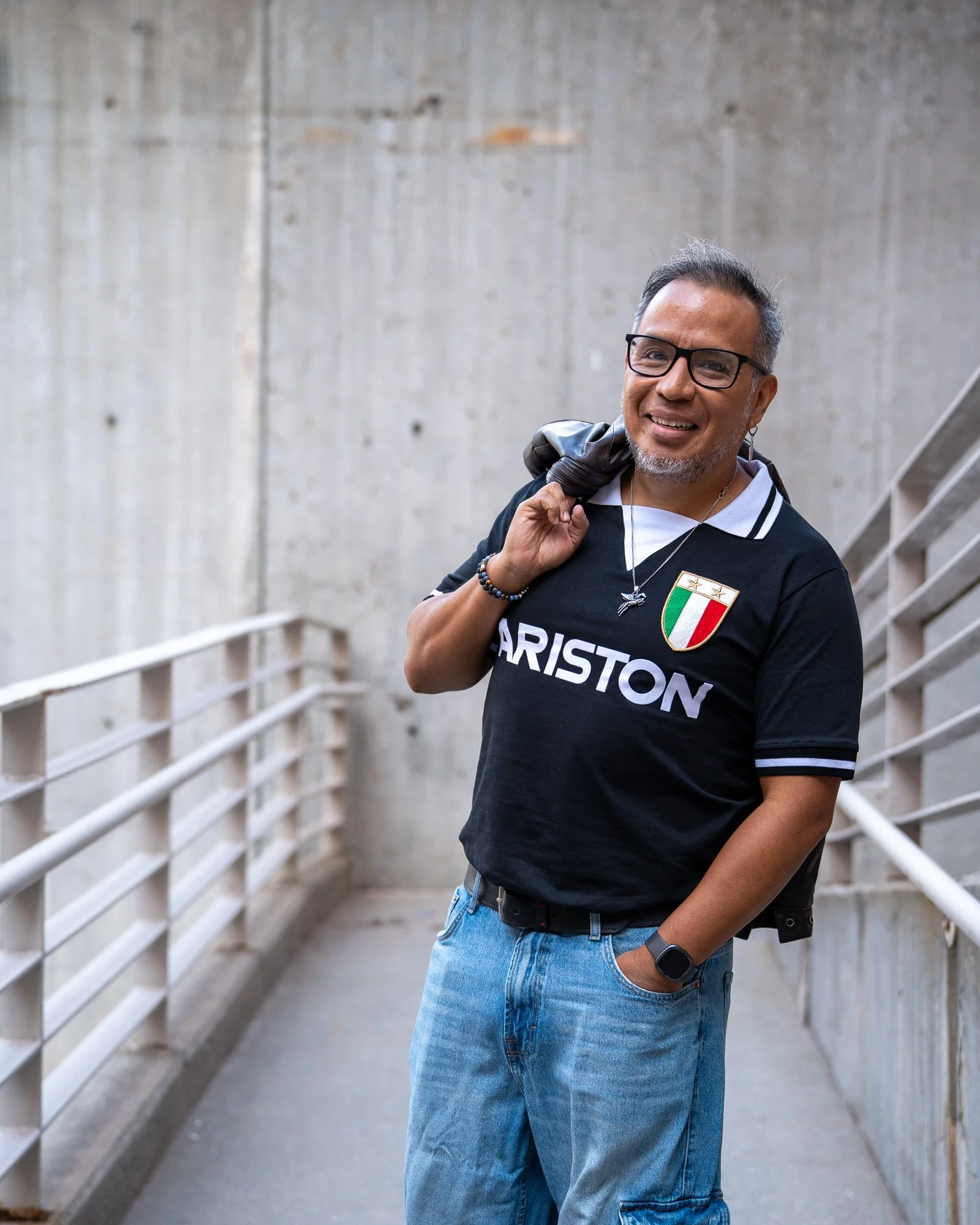 A smiling man with glasses, gray hair, wearing a black t-shirt with the word "PARISTON" and an Italian flag crest, blue jeans, and a wristwatch, standing in front of a concrete wall and metal railing, holding a black jacket over his shoulder.