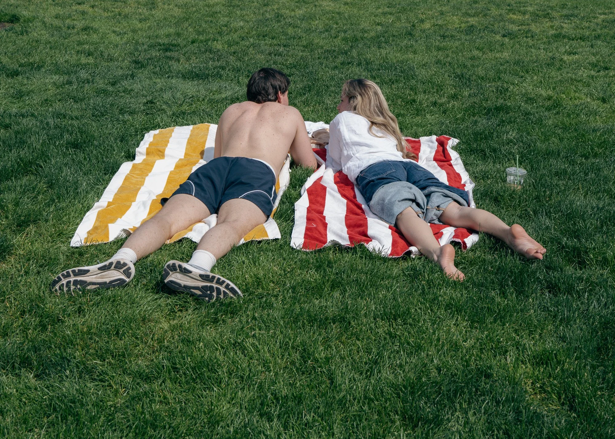 A young man and woman lying on towels on the grass in a park, facing each other and engaging in conversation. The man is shirtless, wearing dark shorts and sneakers, while the woman has long blonde hair, is wearing a white shirt and denim shorts, and