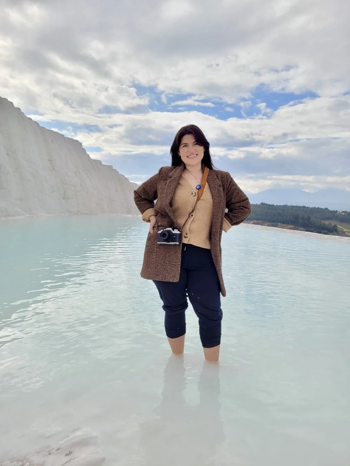 Woman standing in shallow water near white cliff with mountains and cloudy sky in the background, smiling at the camera, wearing a brown coat, beige sweater, black pants, and with a camera hanging around her neck.