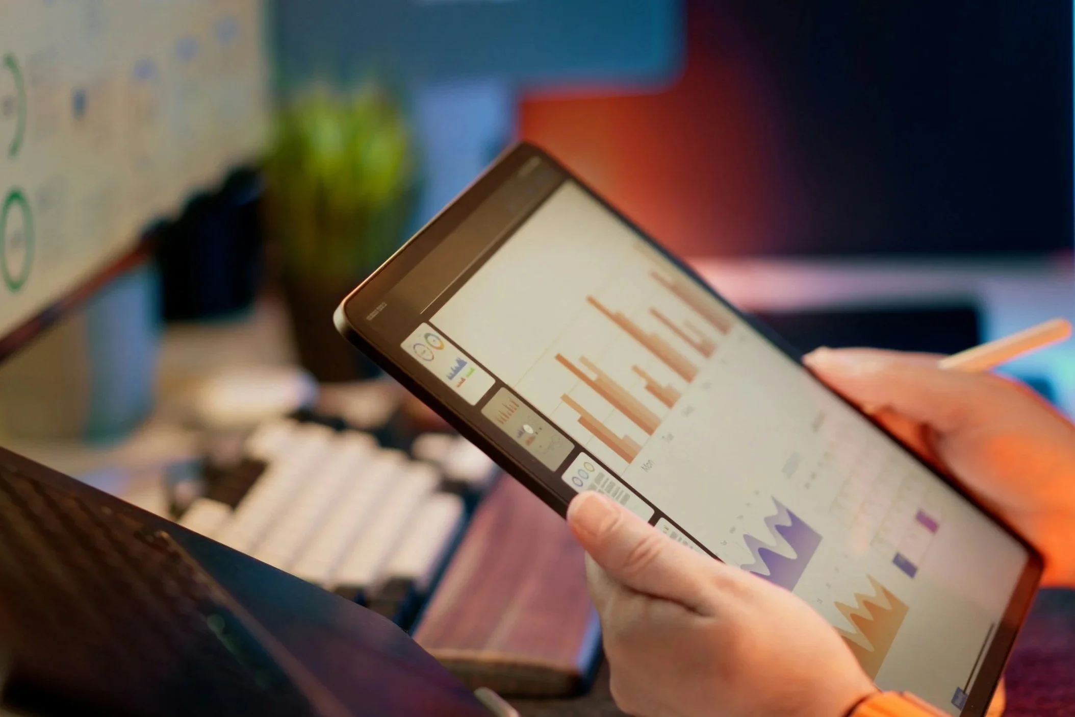 Close-up of a person's hand using a stylus on a tablet displaying a bar graph, with a blurred background of a whiteboard and office environment.