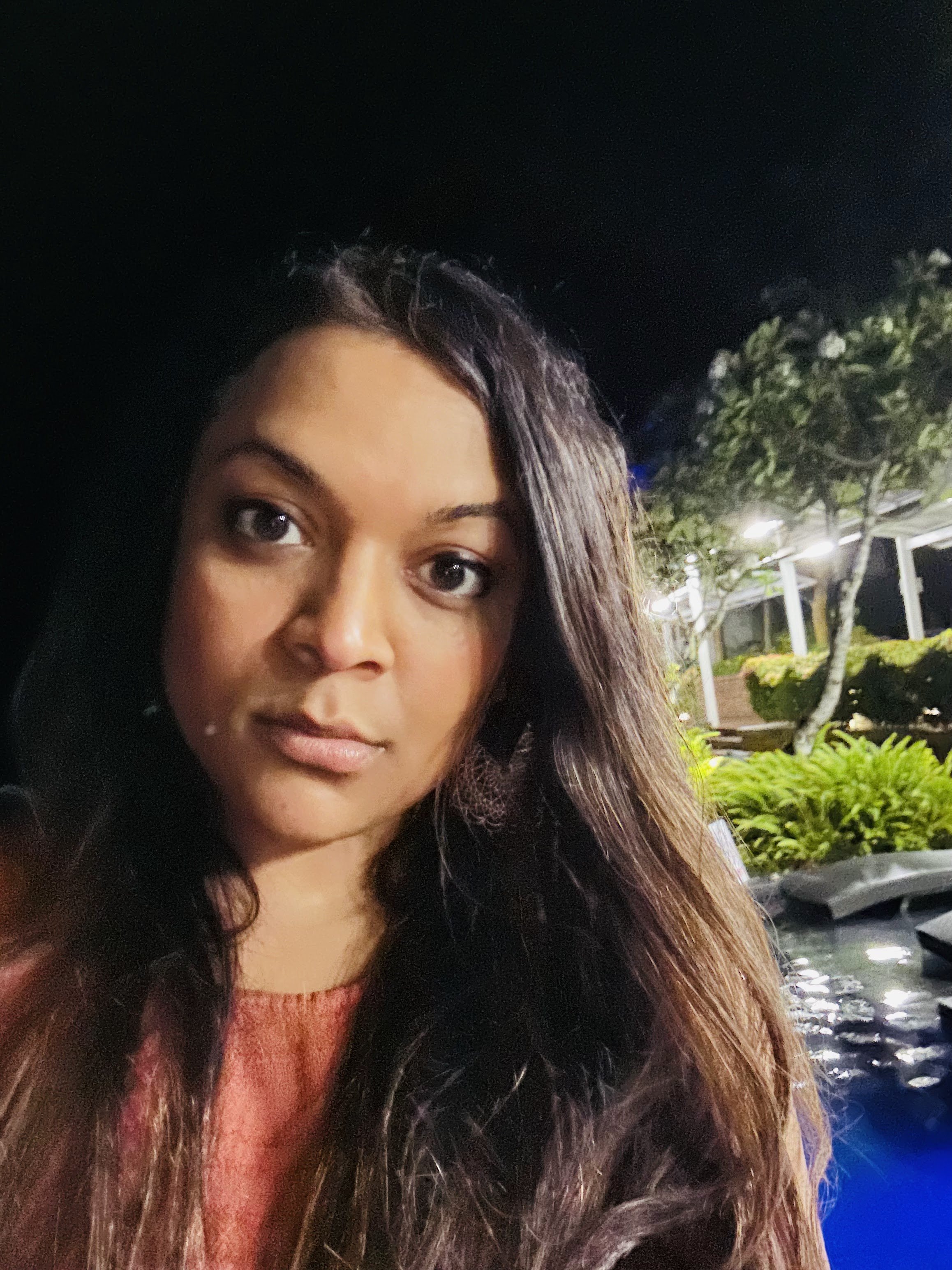 A woman with long dark hair and brown eyes is outdoors at night, with a background of trees, streetlights, and parked cars.