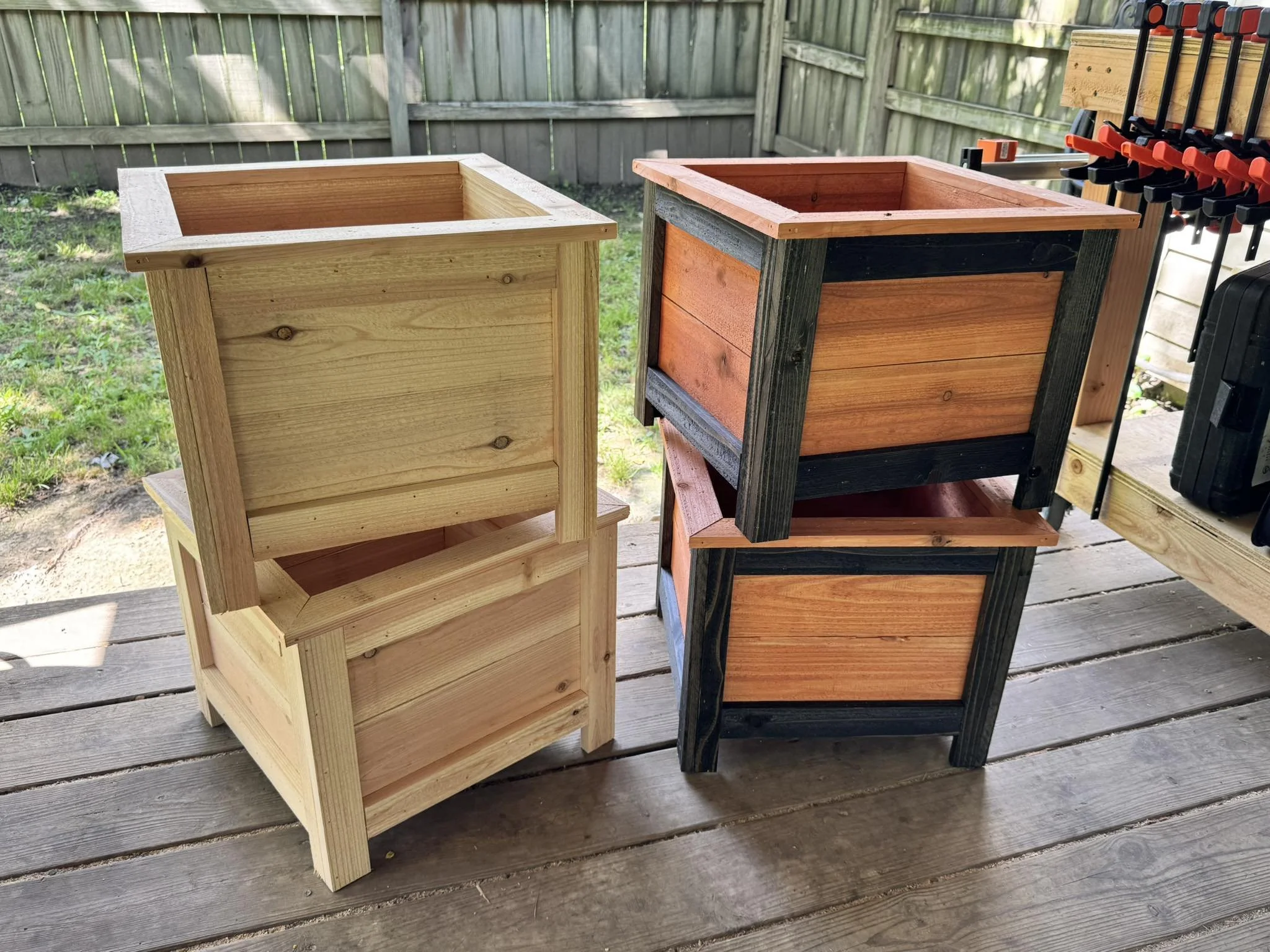 Two empty wooden plant boxes on a wooden porch, with a green fence in the background and tools hanging on a rack to the right.