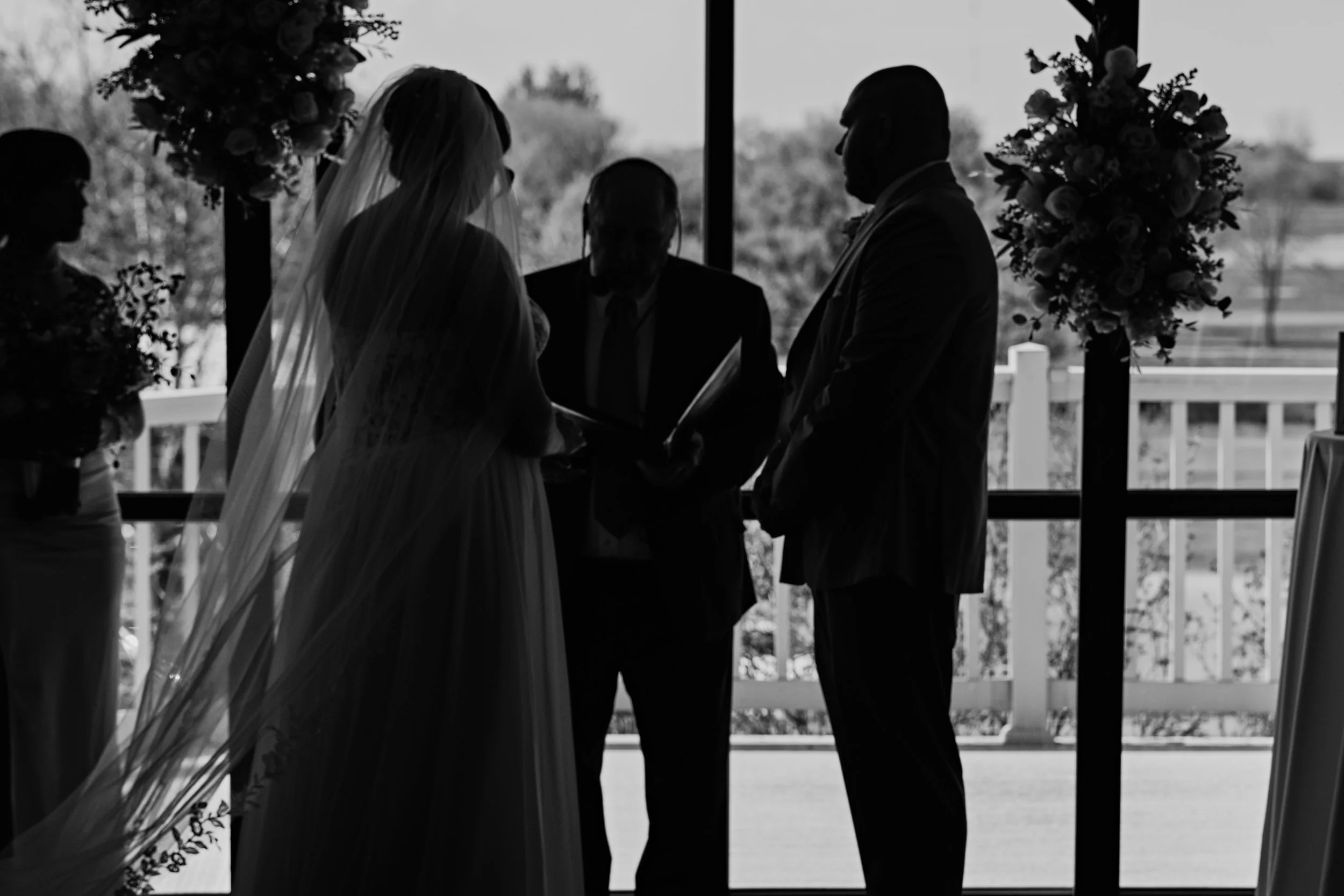 Silhouette of a bride and groom facing each other during a wedding ceremony inside, with a person officiating, near large windows with views of trees outside, and decorated with floral arrangements.