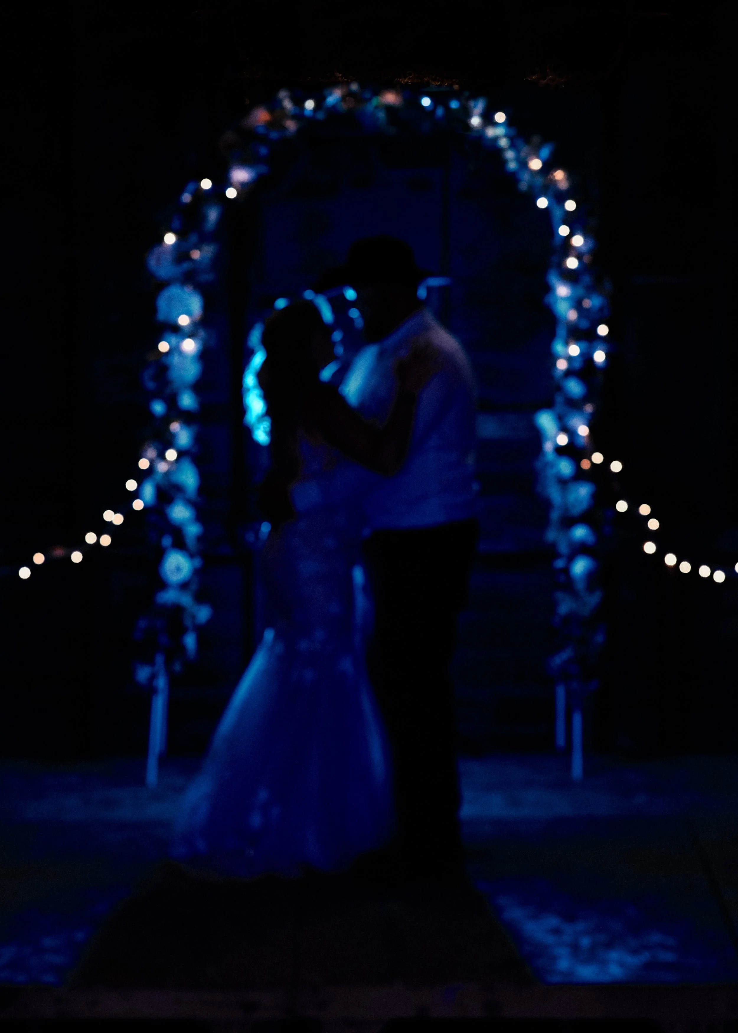 A dark scene with a couple kissing under an archway decorated with blue and white lights, with a long table in the foreground.