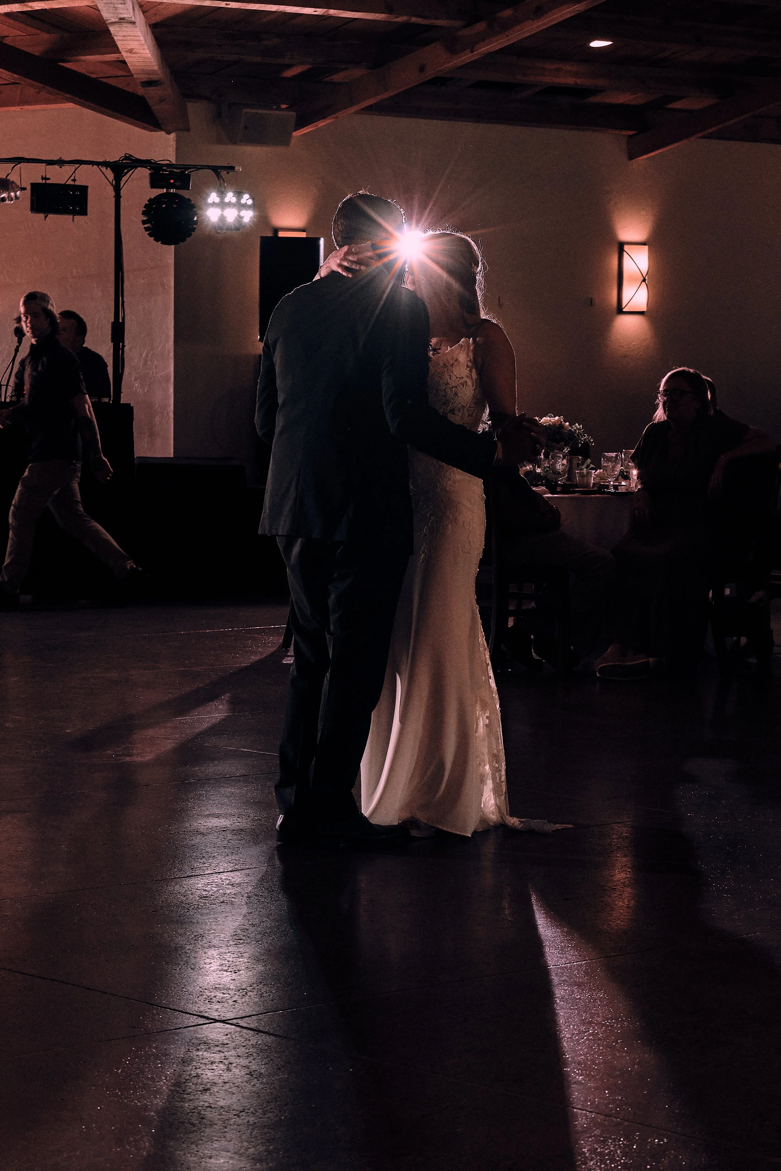 A bride and groom dancing closely at a wedding reception, backlit by a bright light behind them, with guests seated at tables and a DJ or musician visible in the background.
