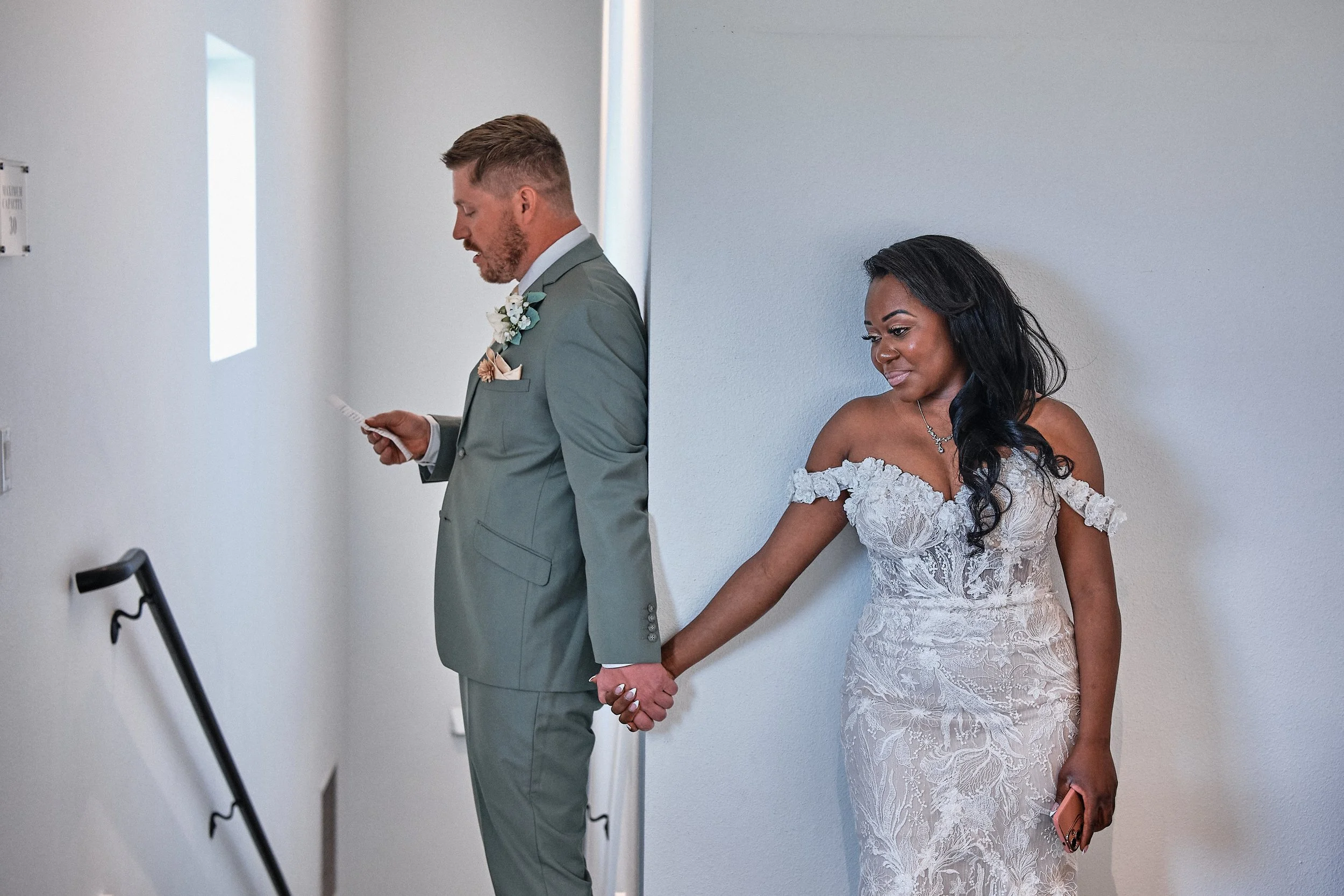 A bride and groom holding hands while standing apart in a wedding ceremony, appearing to be in a hallway or corridor, with both looking down and holding hands behind their backs.