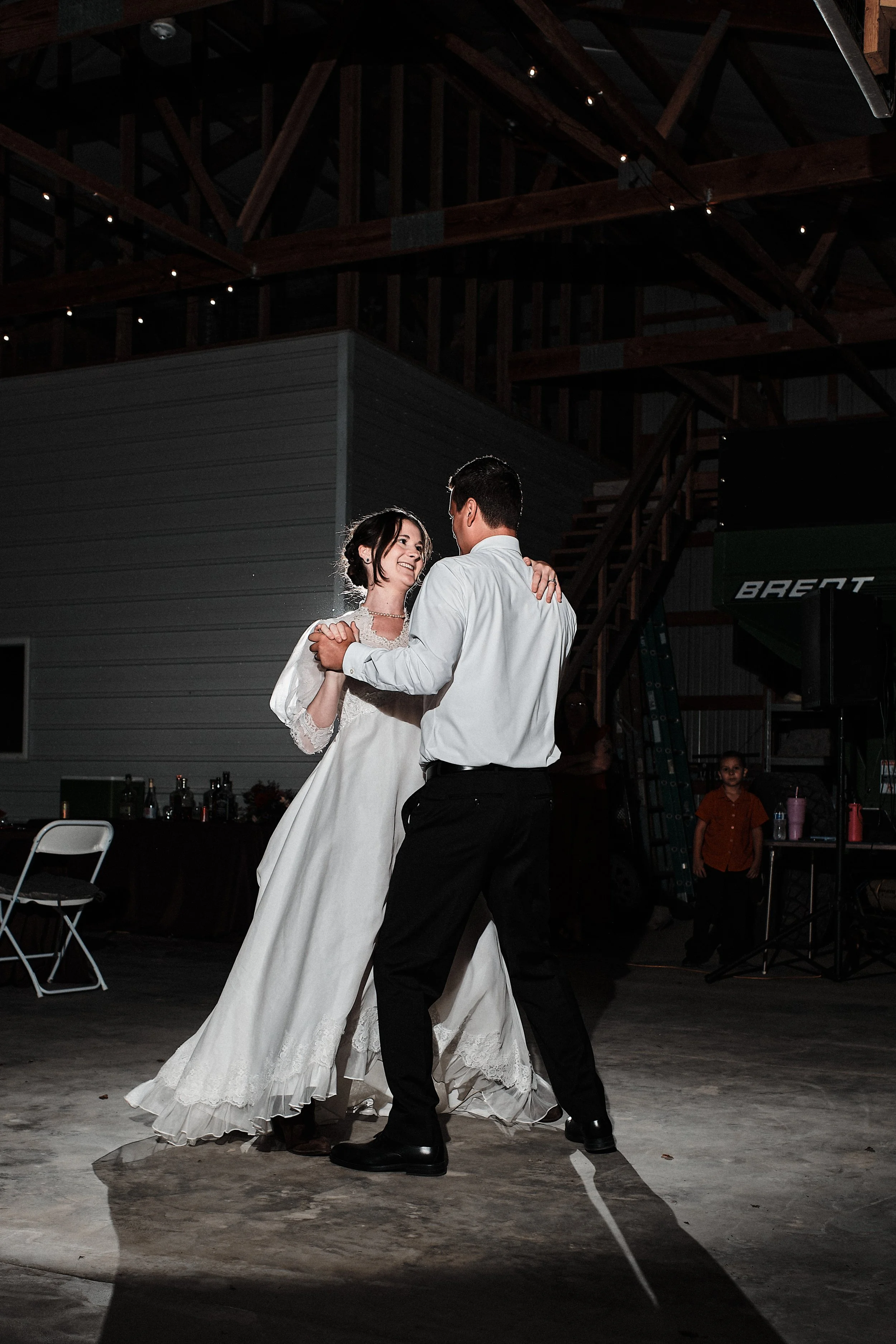 A couple dances together at a wedding reception in a barn-style venue. The bride wears a white wedding gown and the groom wears black pants and a white shirt. They are smiling and looking at each other framed by rustic wooden beams and string lights.