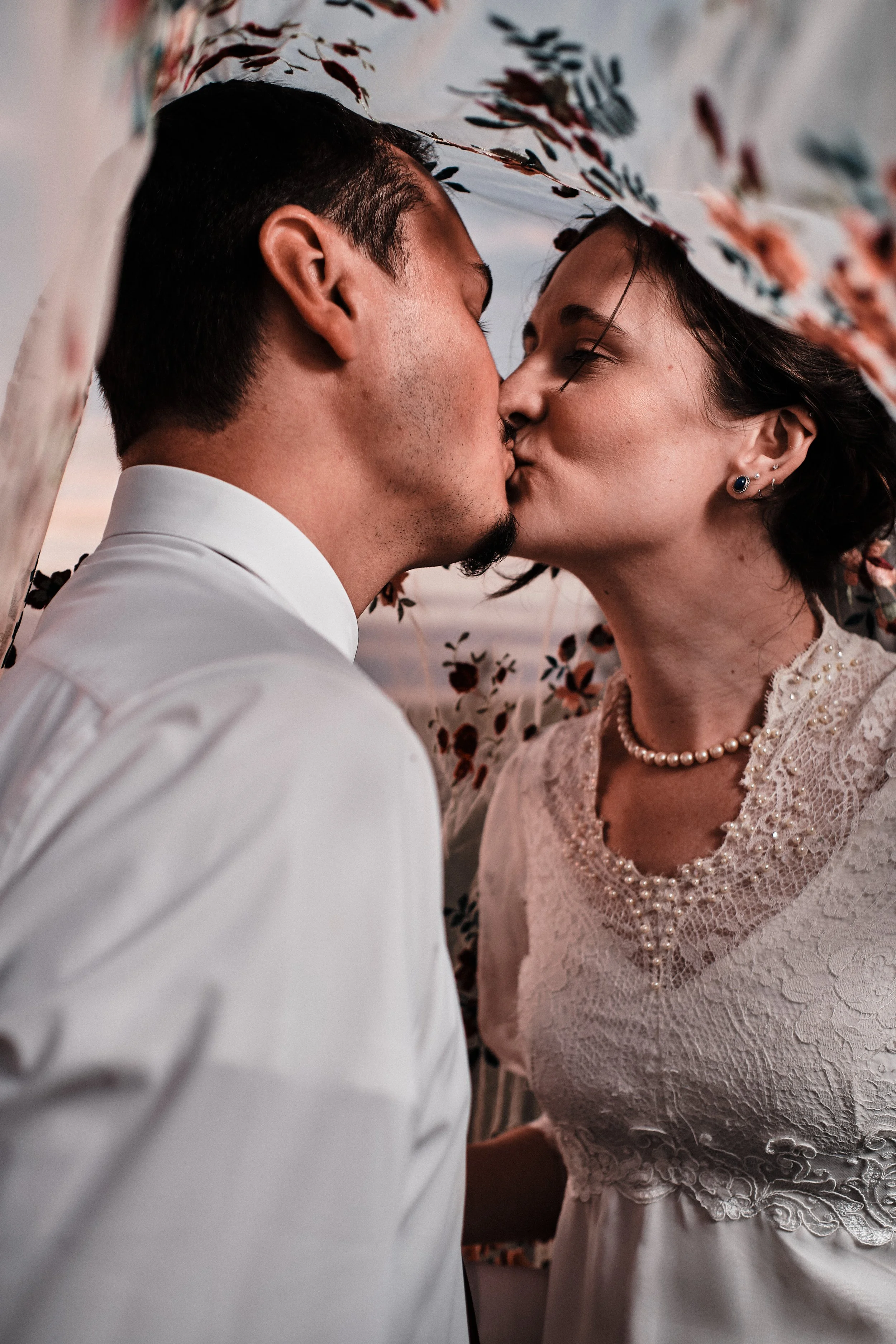 A couple sharing a kiss under a floral canopy during a wedding ceremony. The man is wearing a white shirt, and the woman is dressed in a lace wedding dress with pearl jewelry, including a necklace and earrings.