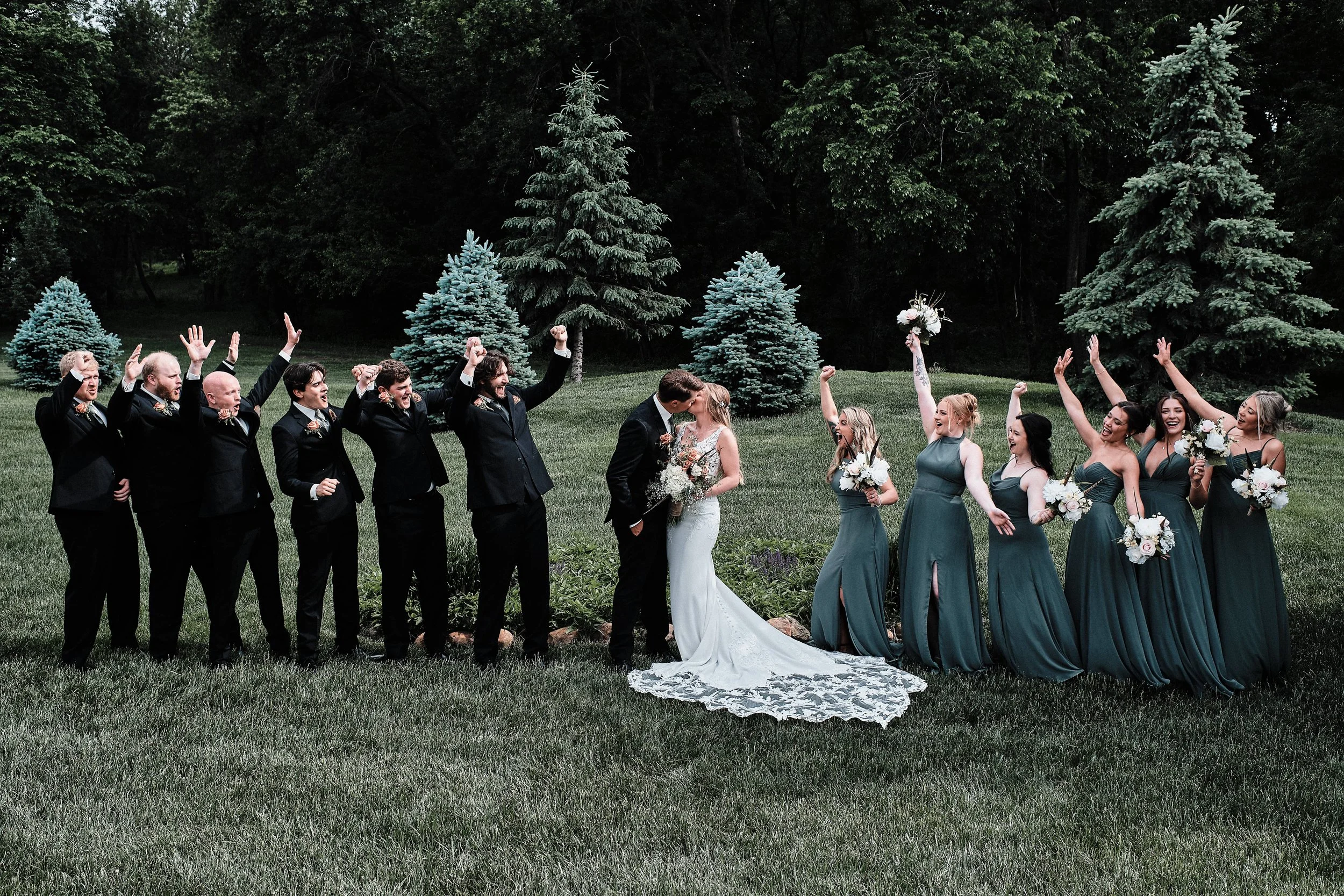 A wedding party outdoors, with the bride and groom in the center, surrounded by their bridesmaids and groomsmen raising their hands in celebration, set against a backdrop of trees and a grassy lawn.