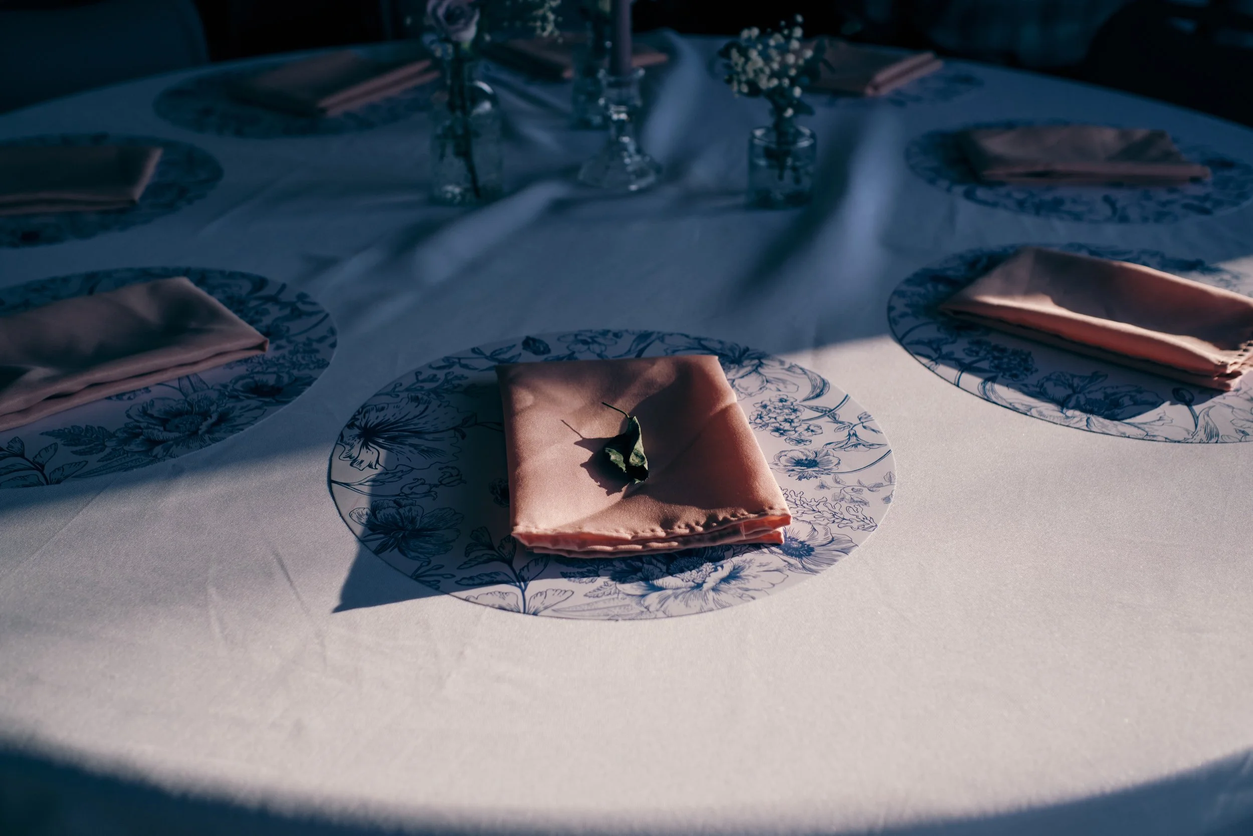 A round dining table with a white tablecloth, set with pink napkins folded on blue and white floral-patterned plates, and decorated with small glass vases holding white flowers, with a single green leaf on one napkin.