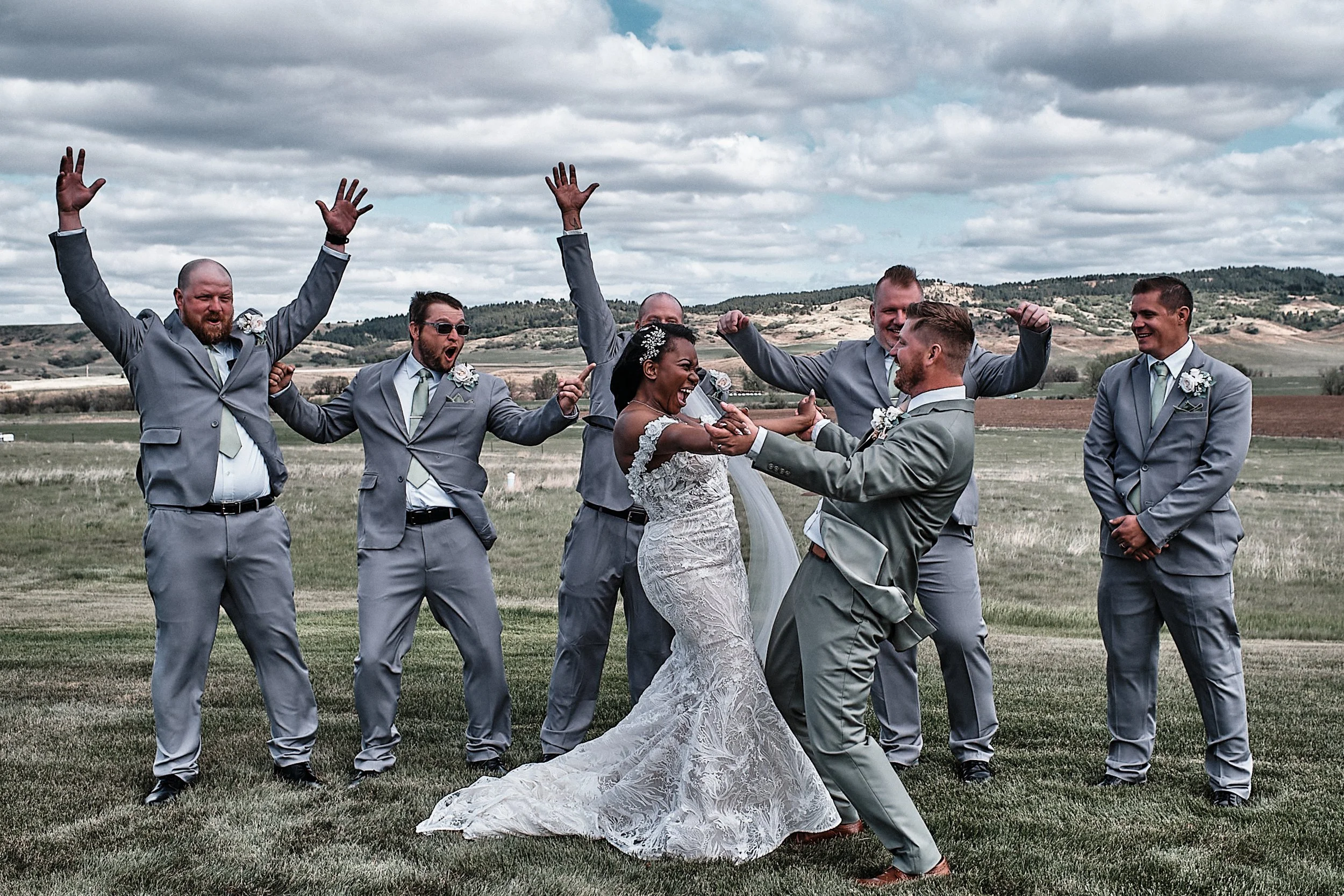 A wedding celebration outdoors with one bride and six grooms, all dressed in formal wedding attire, dancing on a grassy field with mountains and a partly cloudy sky in the background.