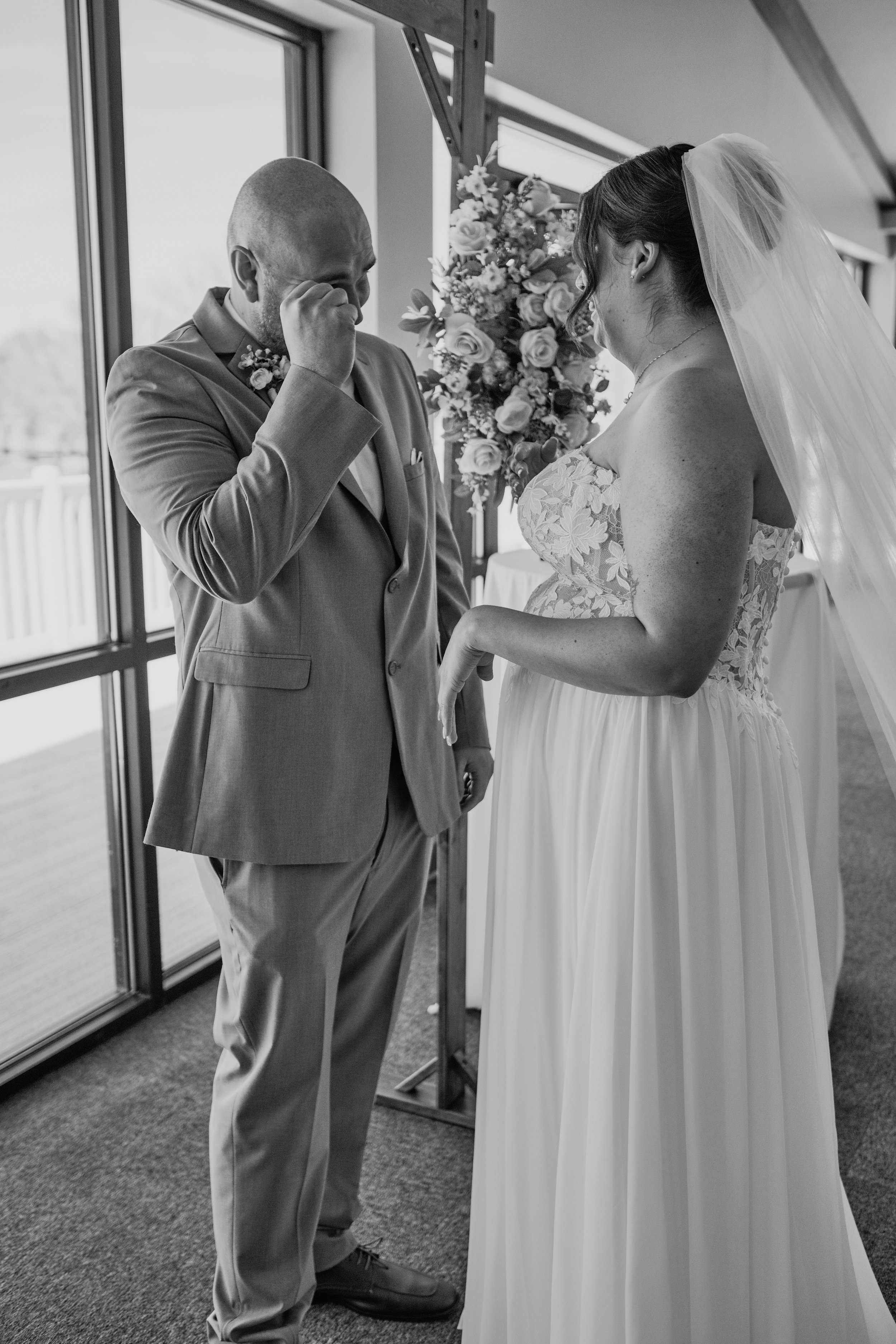 A black and white photo of a wedding ceremony showing a groom tearing up and a bride smiling, holding hands, with a bouquet of flowers nearby, indoors near a large window.