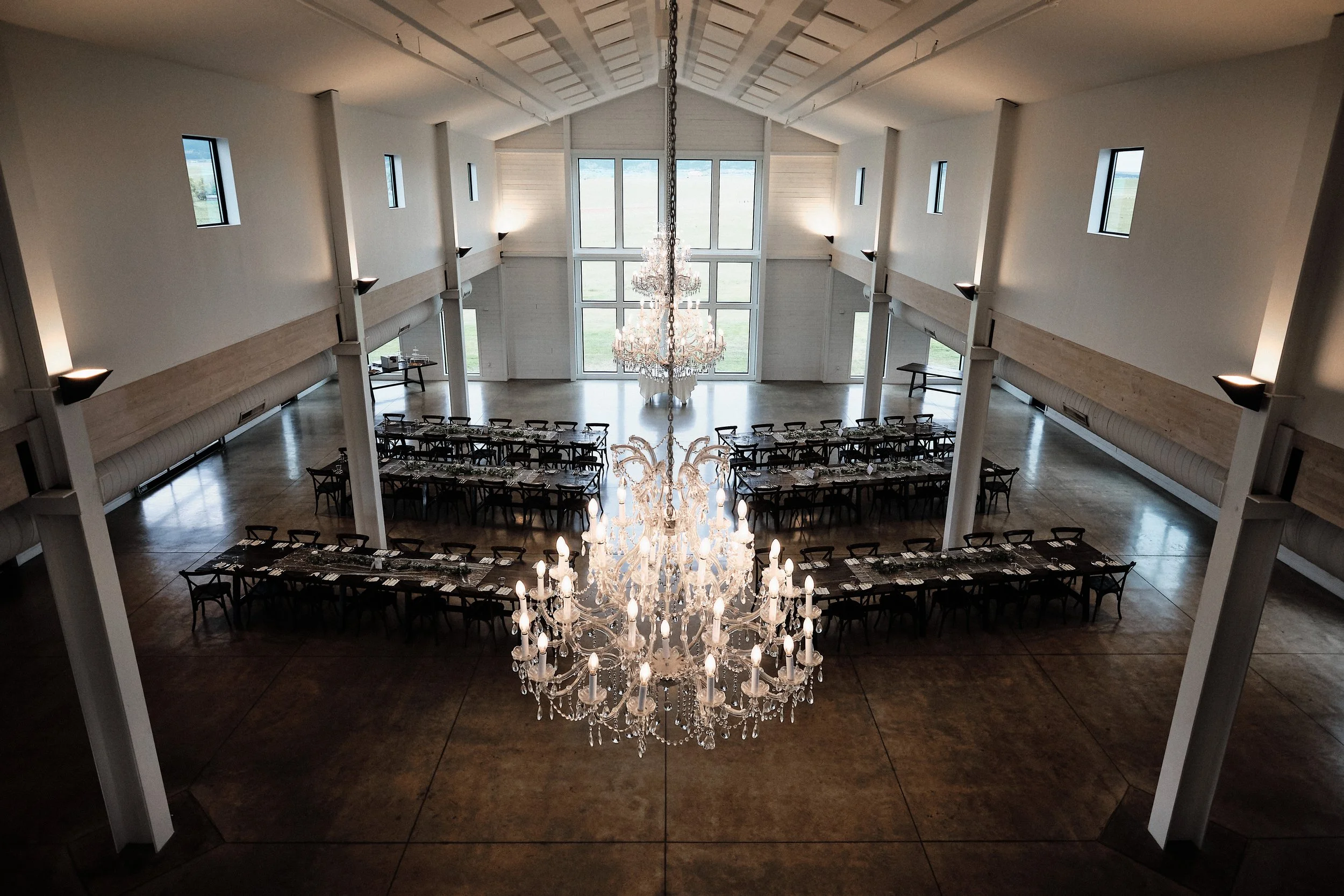 Elegant spacious hall with a large crystal chandelier, long dining tables, multiple windows letting in natural light, and an open view outside.