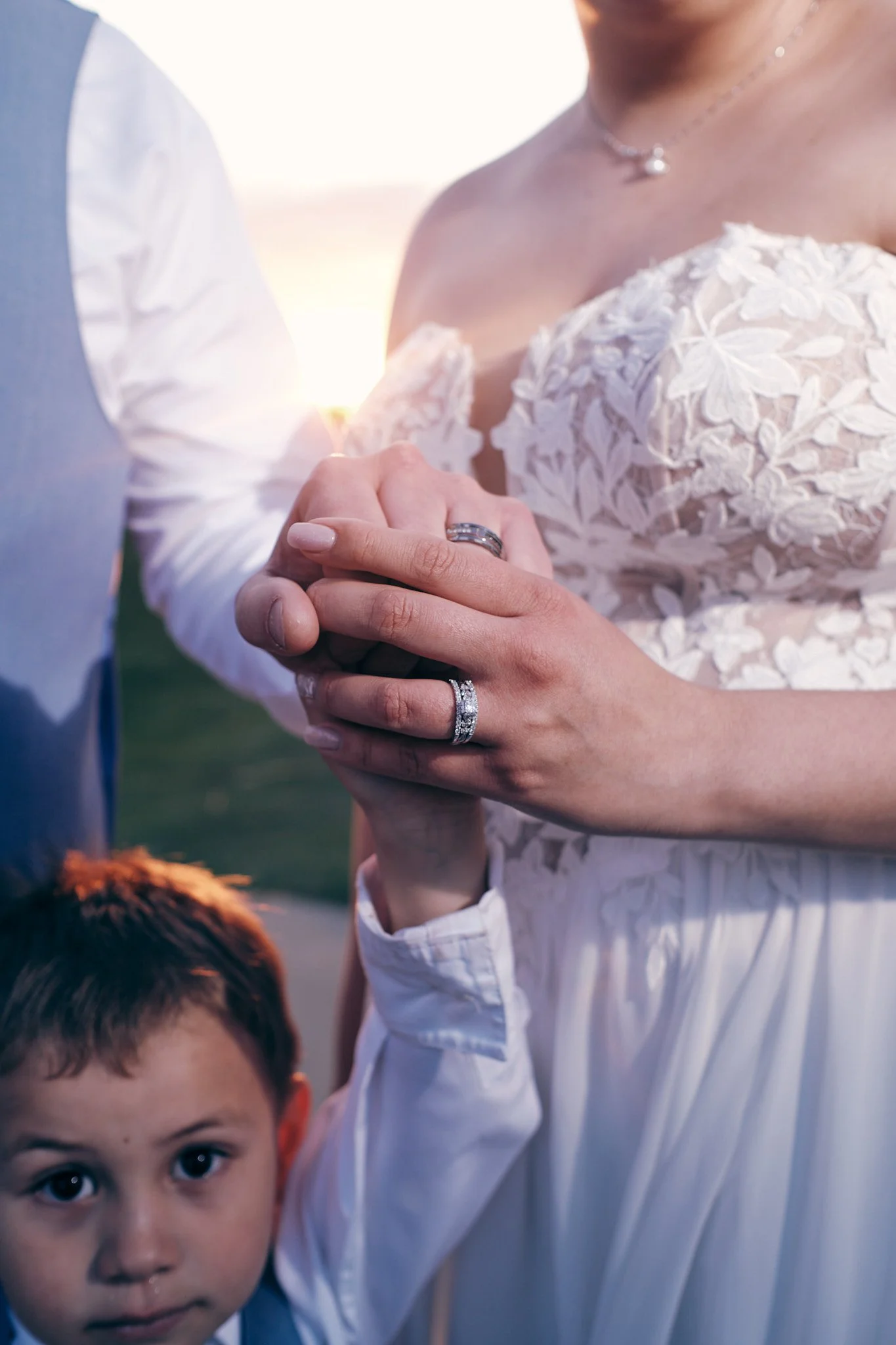 Close-up of a couple holding hands, with the woman wearing wedding rings, and a child with brown hair and big blue eyes looking at the camera in the foreground. The background appears to be outdoors during sunset.