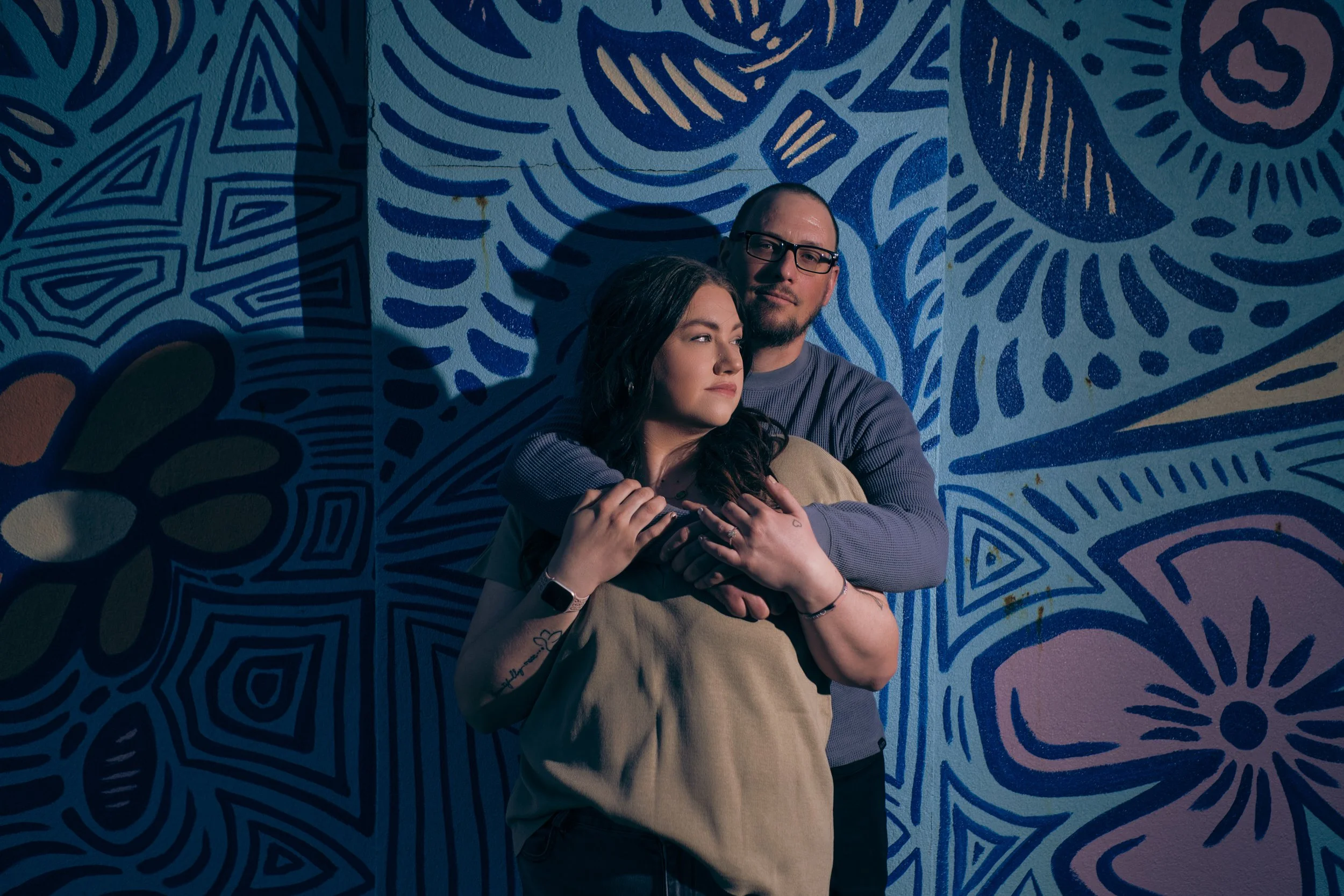 A couple standing in front of a vibrant blue abstract mural, with the woman embracing the man from the front and both looking thoughtful.