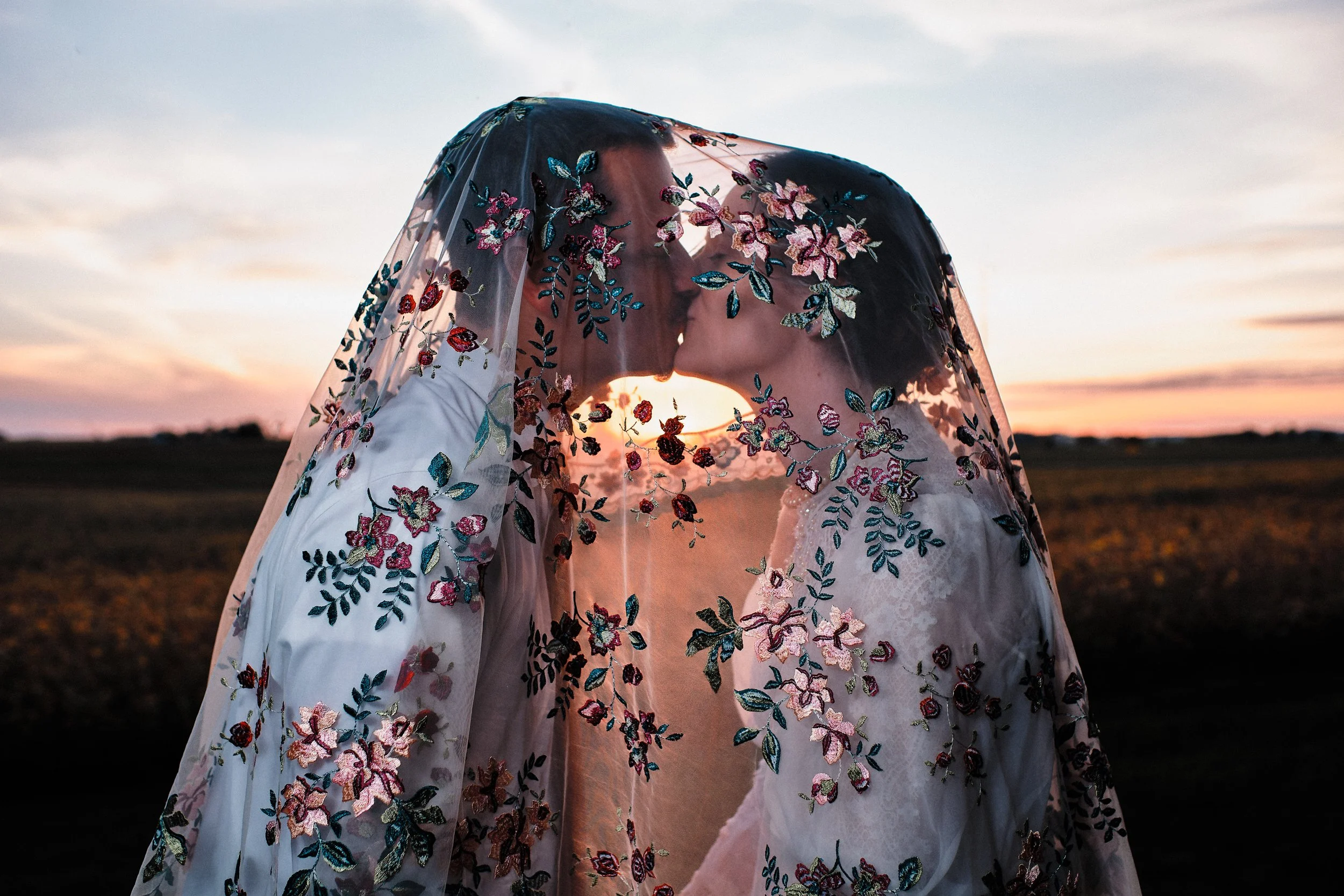 A couple kissing outdoors at sunset, wrapped in a sheer, embroidered fabric with floral designs.