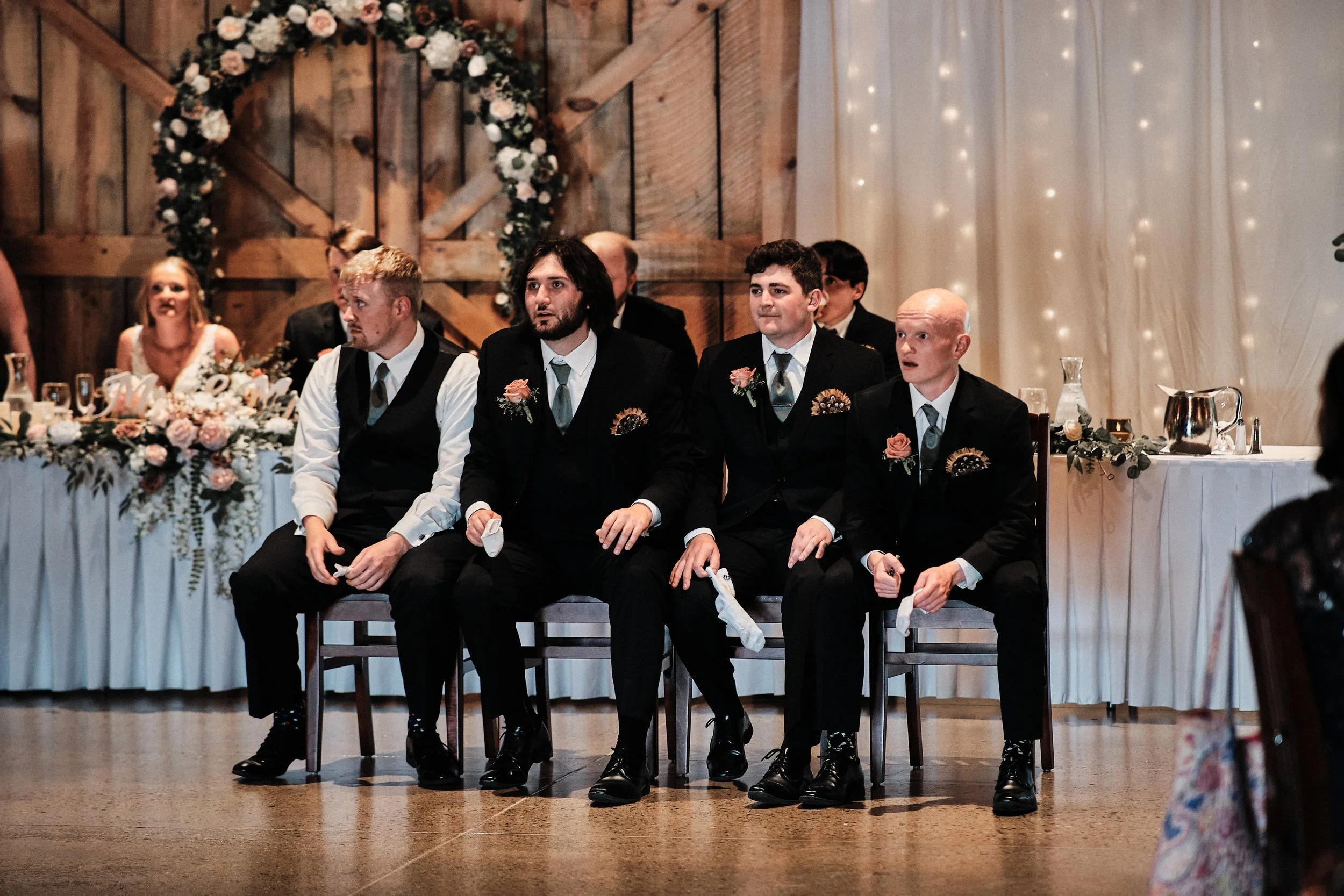 Four men dressed in formal suits sitting at a wedding reception. They have boutonnieres on their lapels and are holding socks. The background features a decorated wedding arch with flowers and fairy lights on a wooden wall.