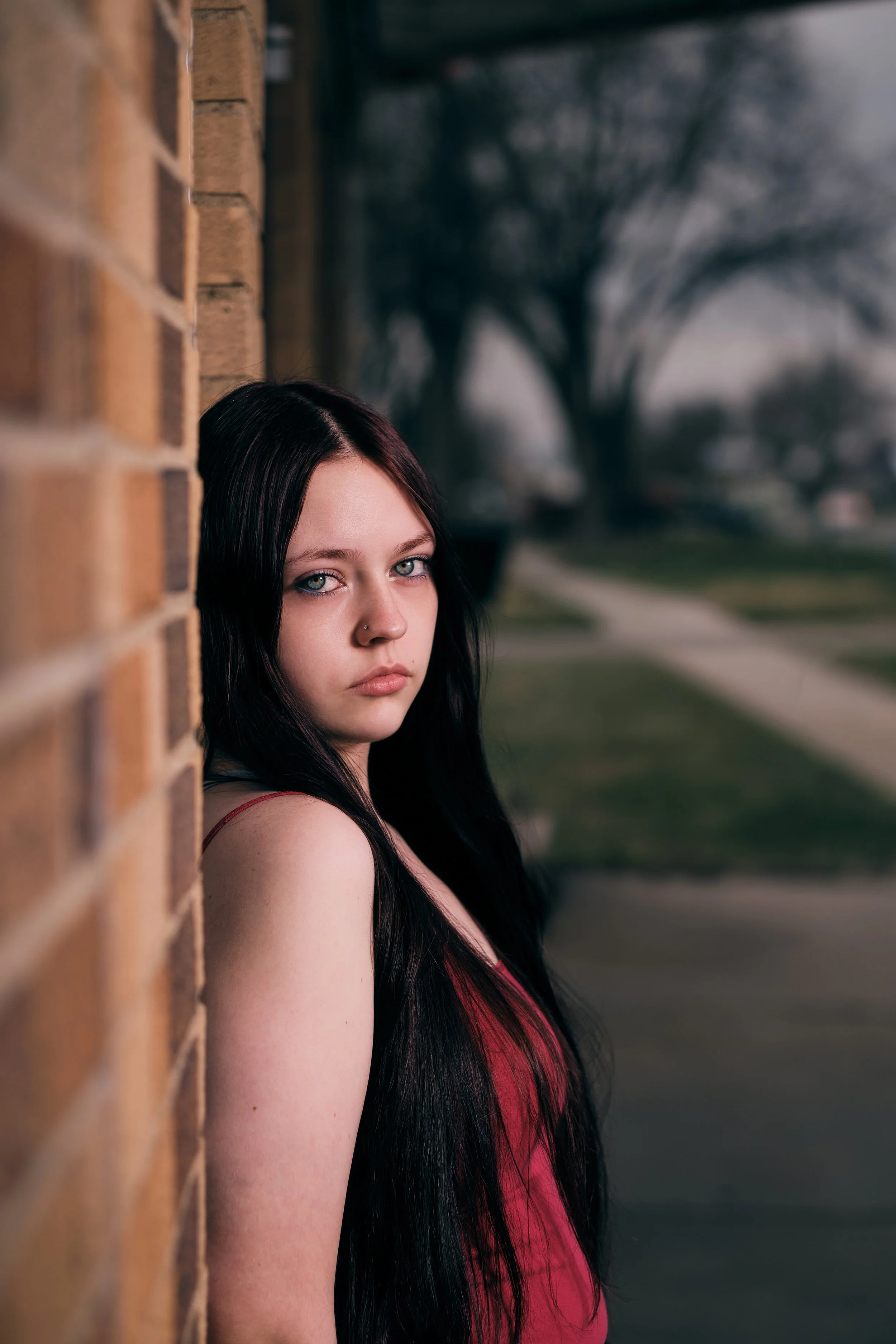 A young woman with long black hair, wearing a red dress, is standing outdoors next to a brick wall, with a blurred background of trees and a lawn.