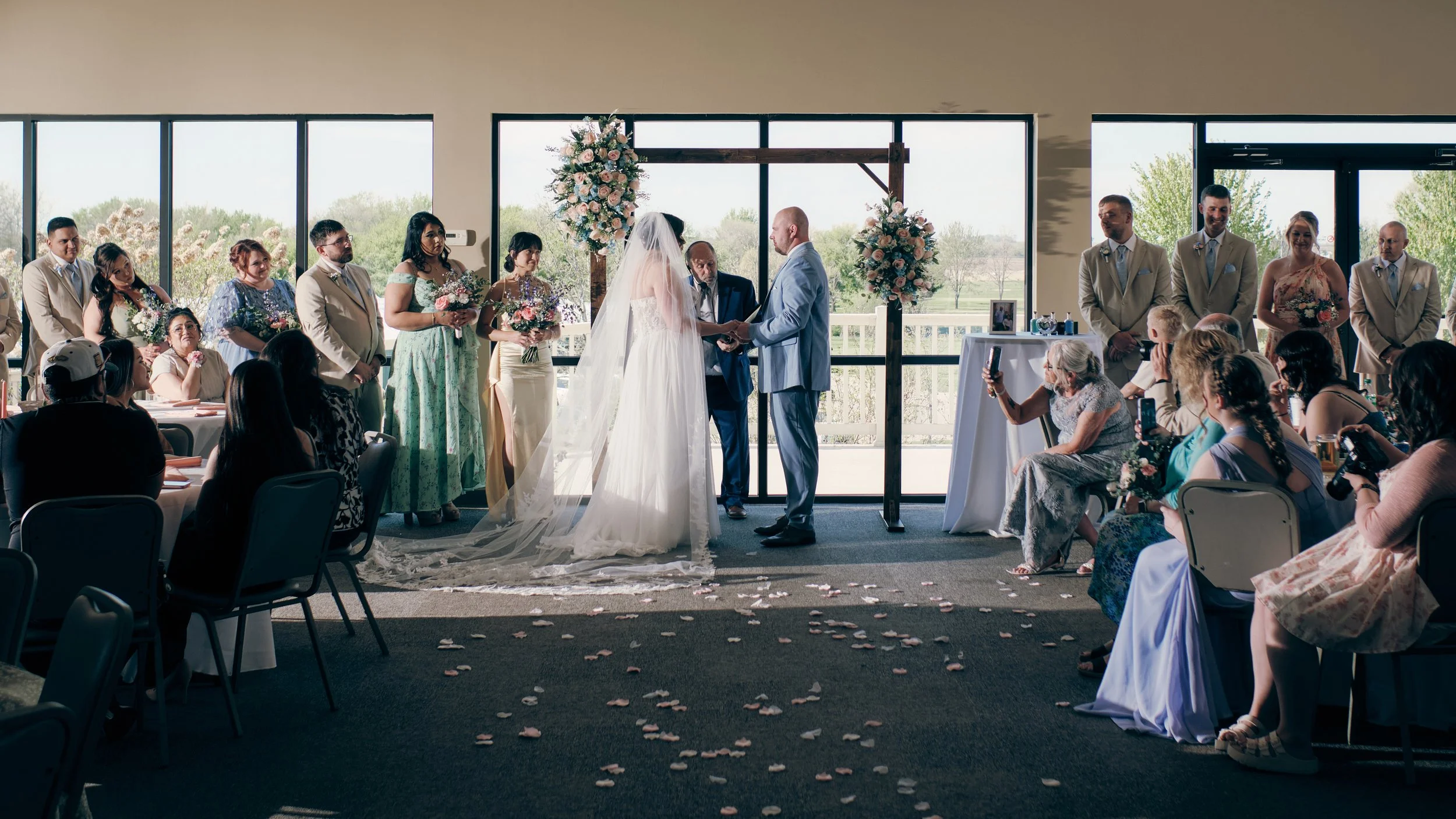 A wedding ceremony taking place indoors with a bride and groom holding hands in front of an officiant, surrounded by bridesmaids and groomsmen. Guests are seated, some taking photos, flowers and petals decorate the aisle, and large windows behind the