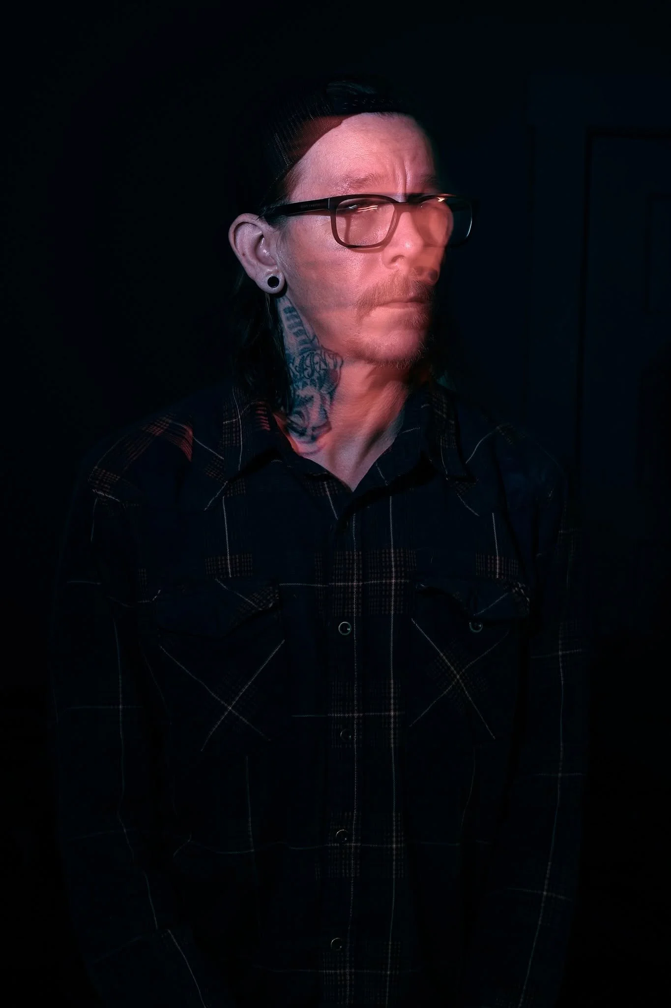 A person with long dark hair wearing glasses, a black plaid shirt, and visible tattoos on their neck and face, standing against a dark background