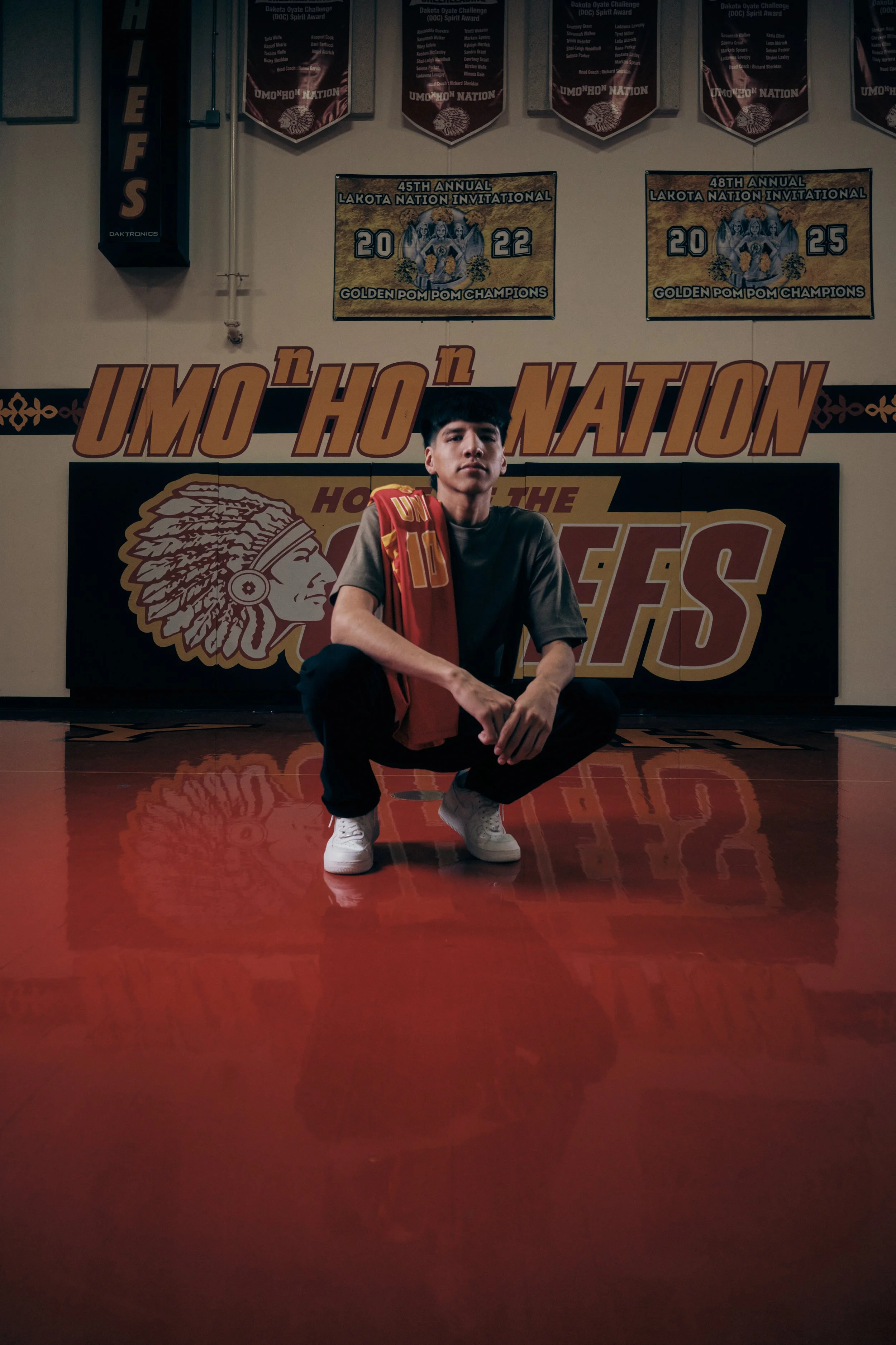Young man squatting on a gym floor, wearing a gray t-shirt with red and yellow cloth draped over his shoulder, in a gym decorated with banners and posters, including championship posters for the Lakota Nation Invitational.