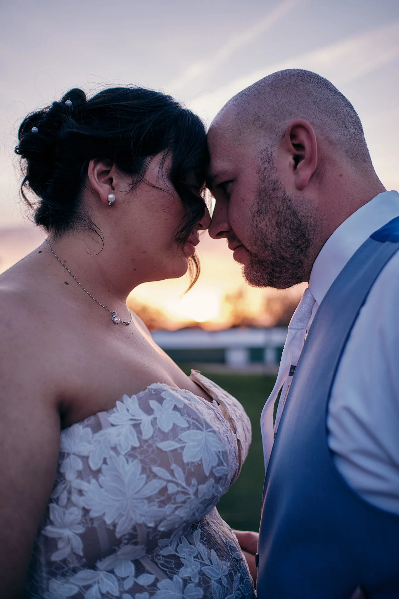 A bride and groom are standing close with their foreheads touching during sunset. The bride is wearing a strapless lace wedding dress with floral patterns, pearl earrings, and a matching necklace. The groom is in a light blue suit with a white shirt 
