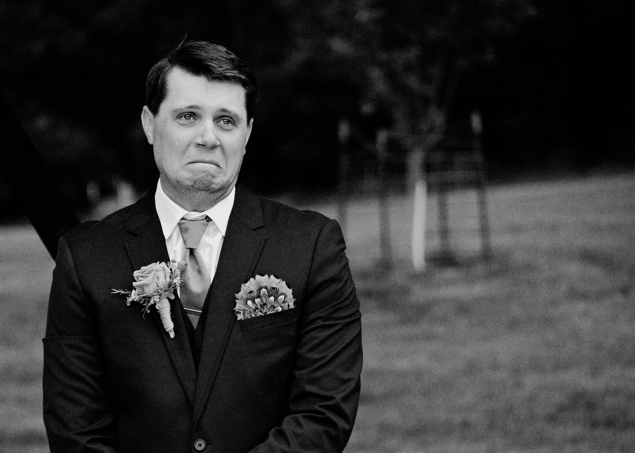 Black and white photo of a man with dark hair dressed in a suit, wearing a tie, with flowers pinned to his lapel, outdoors with a blurred background.