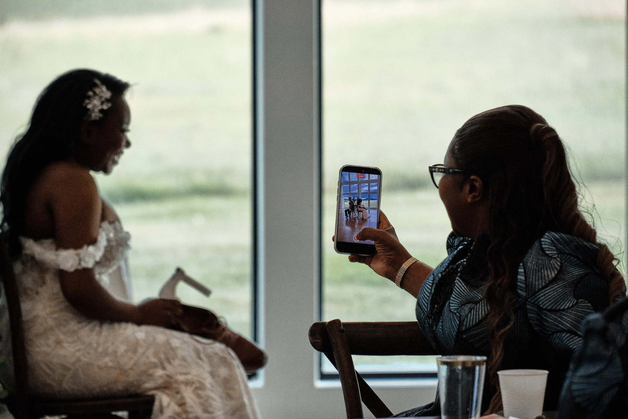 A woman taking a photo of a young girl dressed in a white dress with floral details, sitting in front of a large window with a blurred outdoor view.